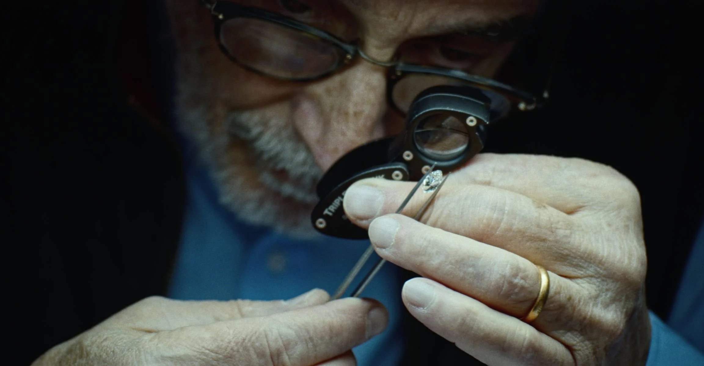 An older man with glasses using a magnifying glass to carefully examine a small watch part.