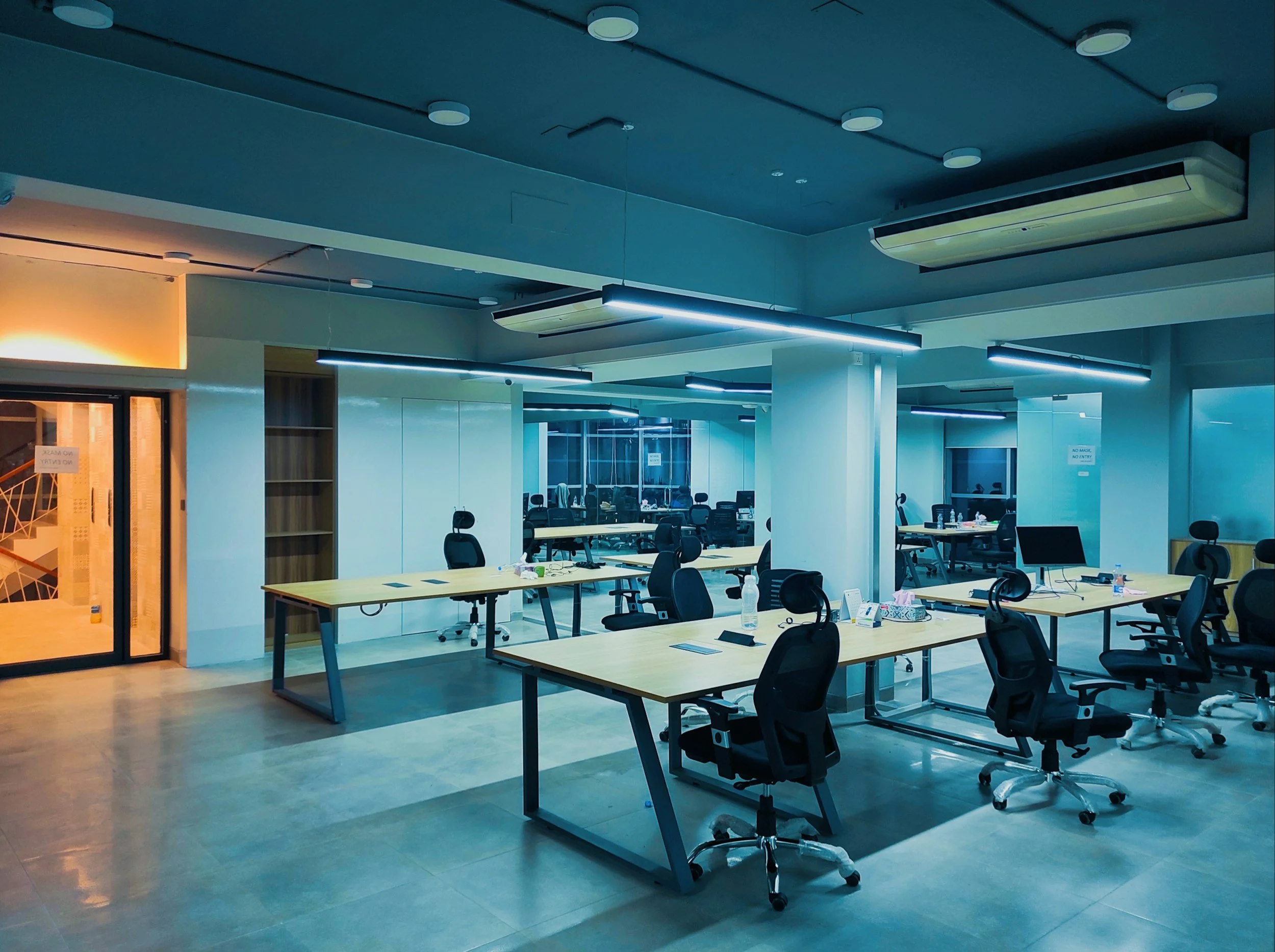 Empty modern office with desks, chairs, and water bottles, illuminated by overhead lights in a spacious environment.