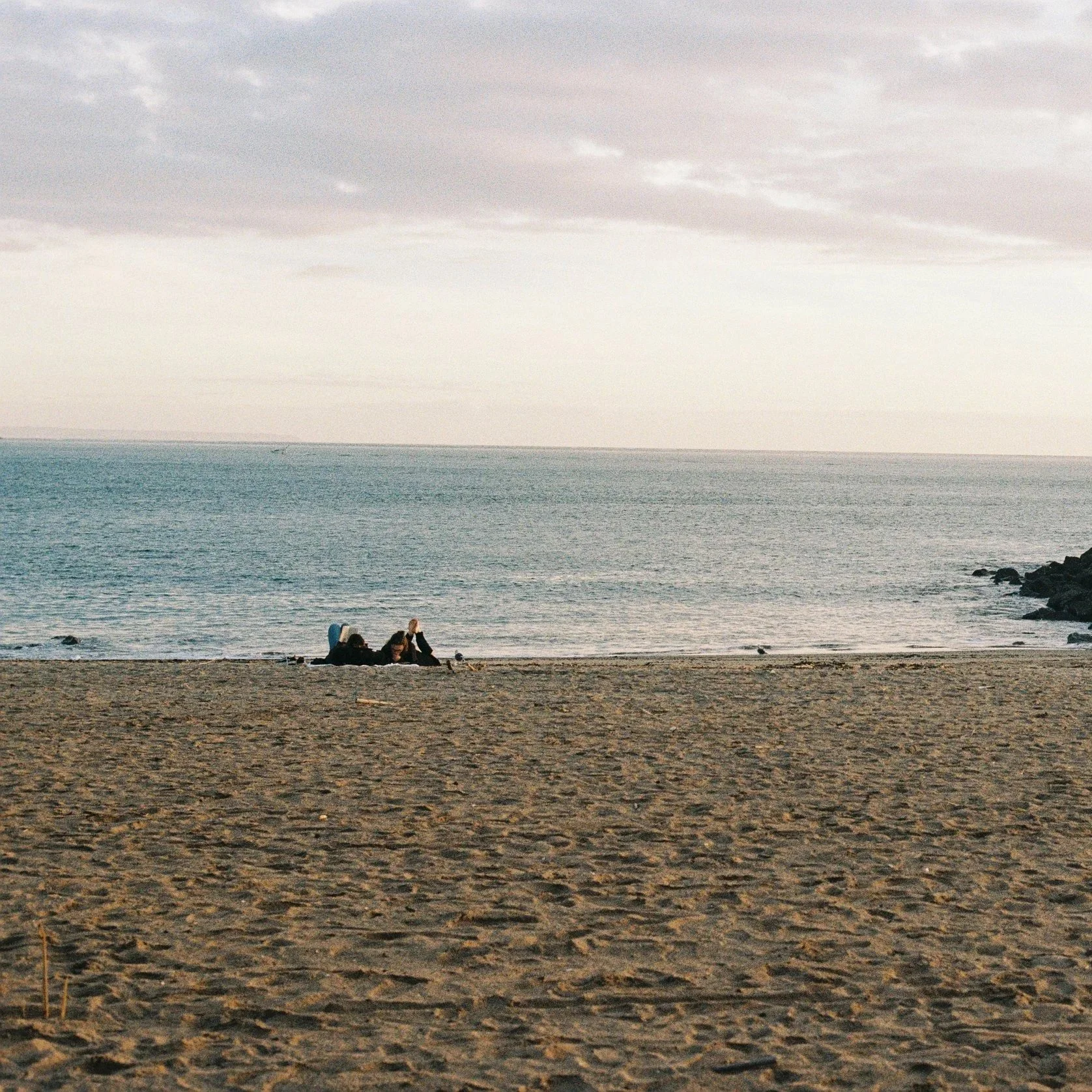 A beach scene with three people sitting on the sand near the water's edge, with the ocean extending into the horizon and a cloudy sky above.