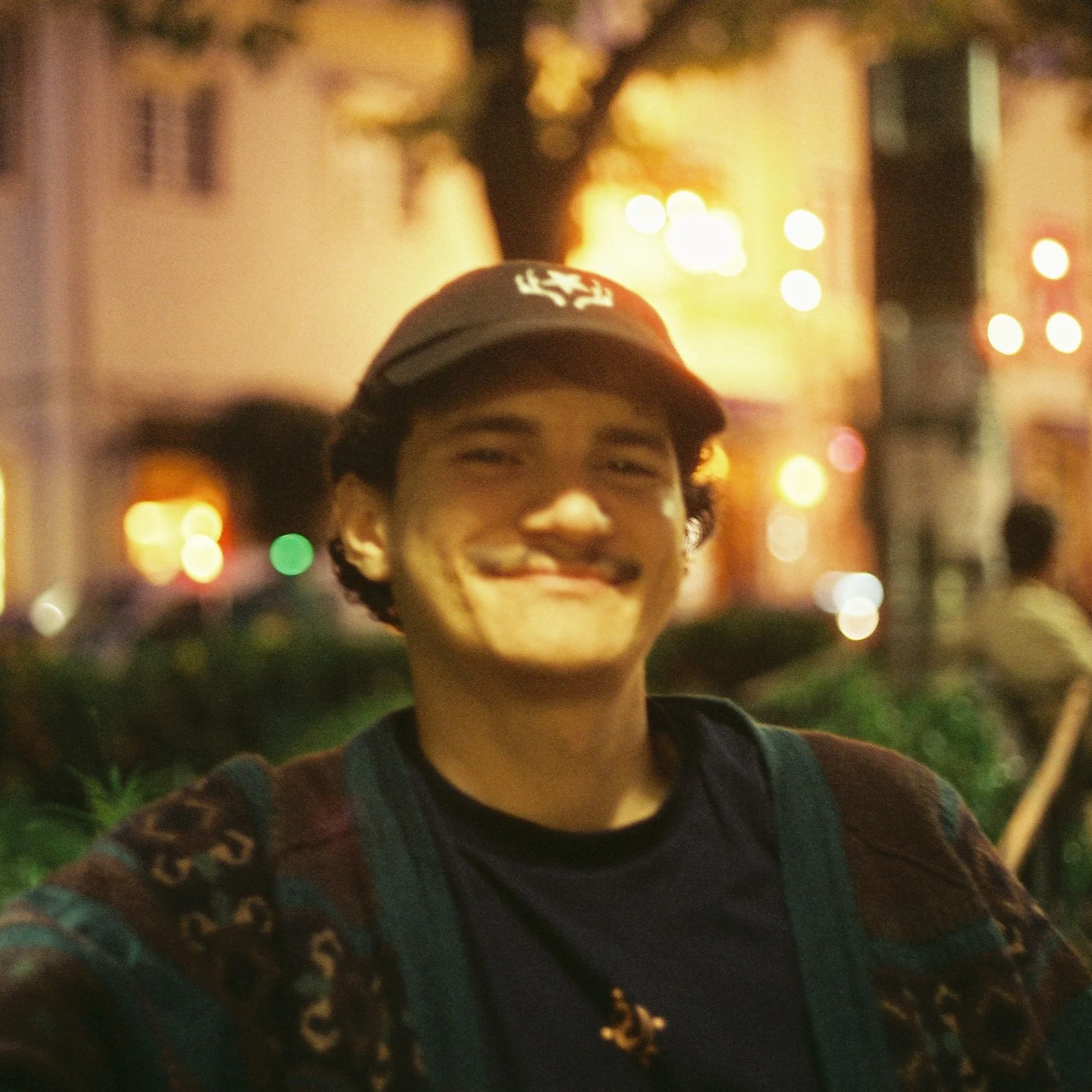 A young person smiling at night with blurred city lights in the background, wearing a baseball cap and a patterned sweater.