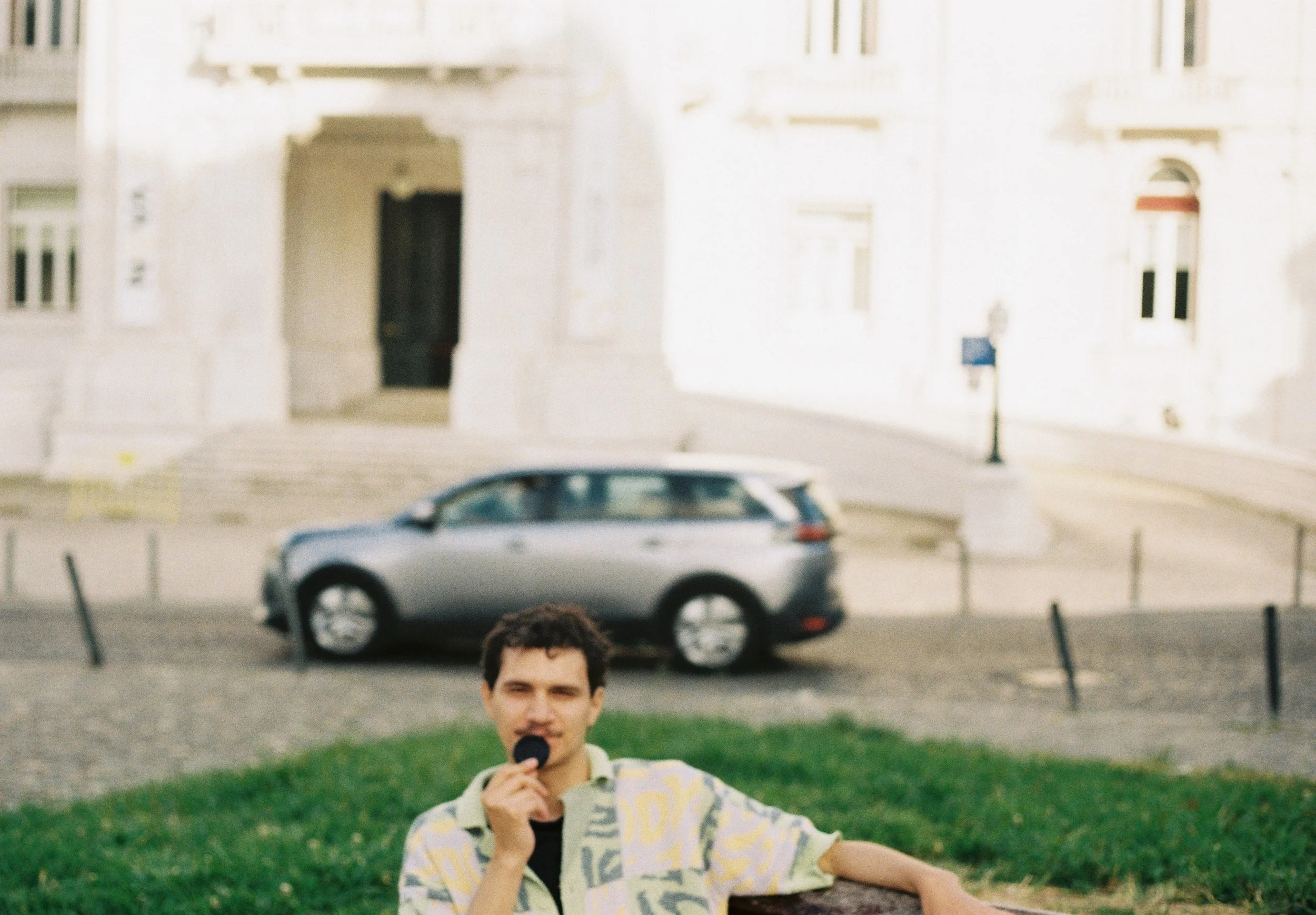 A young man with dark curly hair wearing a yellow patterned shirt, holding a microphone, sitting on a stone ledge in a park with green grass, with an indistinct family or couple in the background and modern buildings.