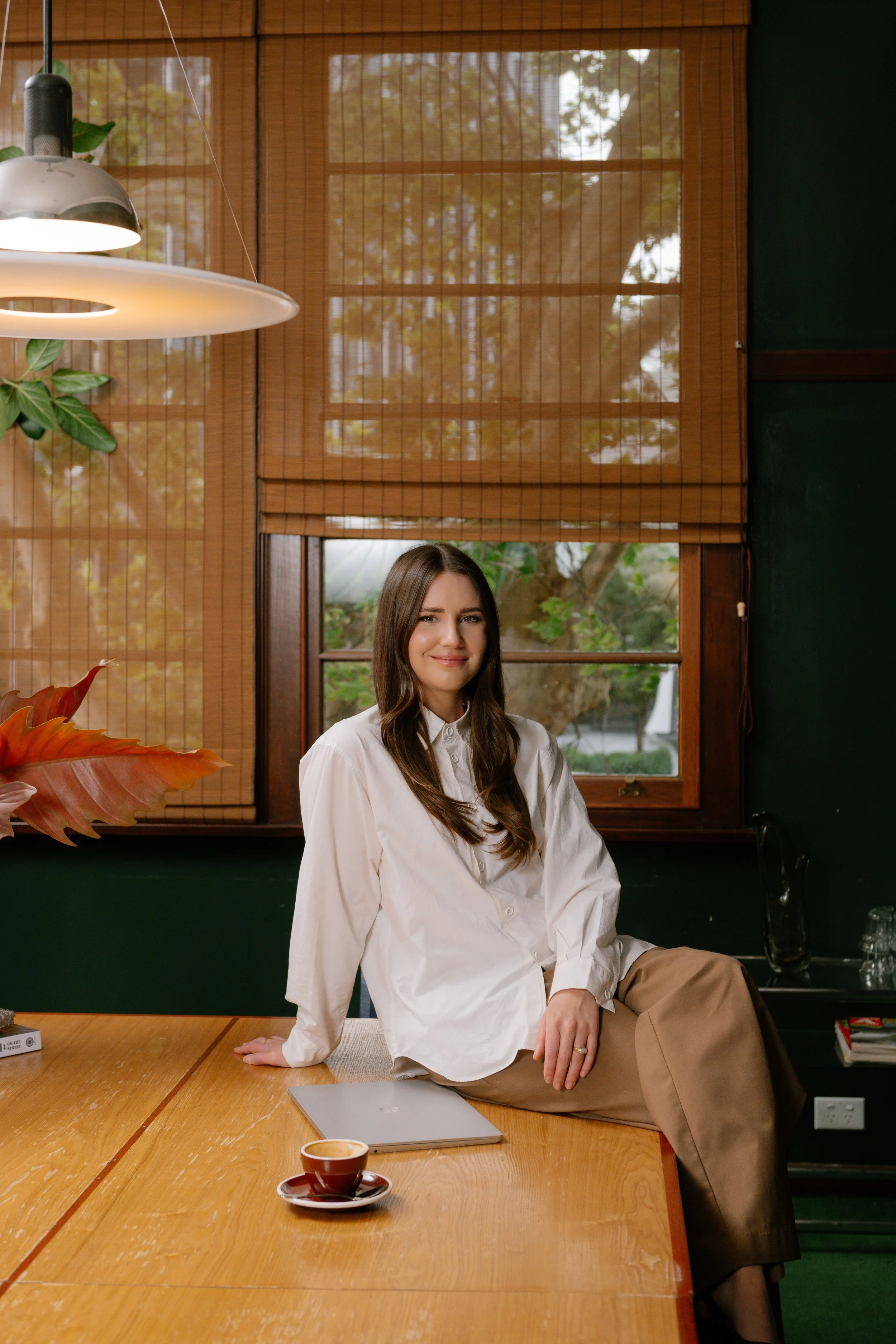 A woman with long brown hair wearing a white shirt sitting on a wooden table with a laptop and a cup of coffee, with a window showing trees outside in the background.