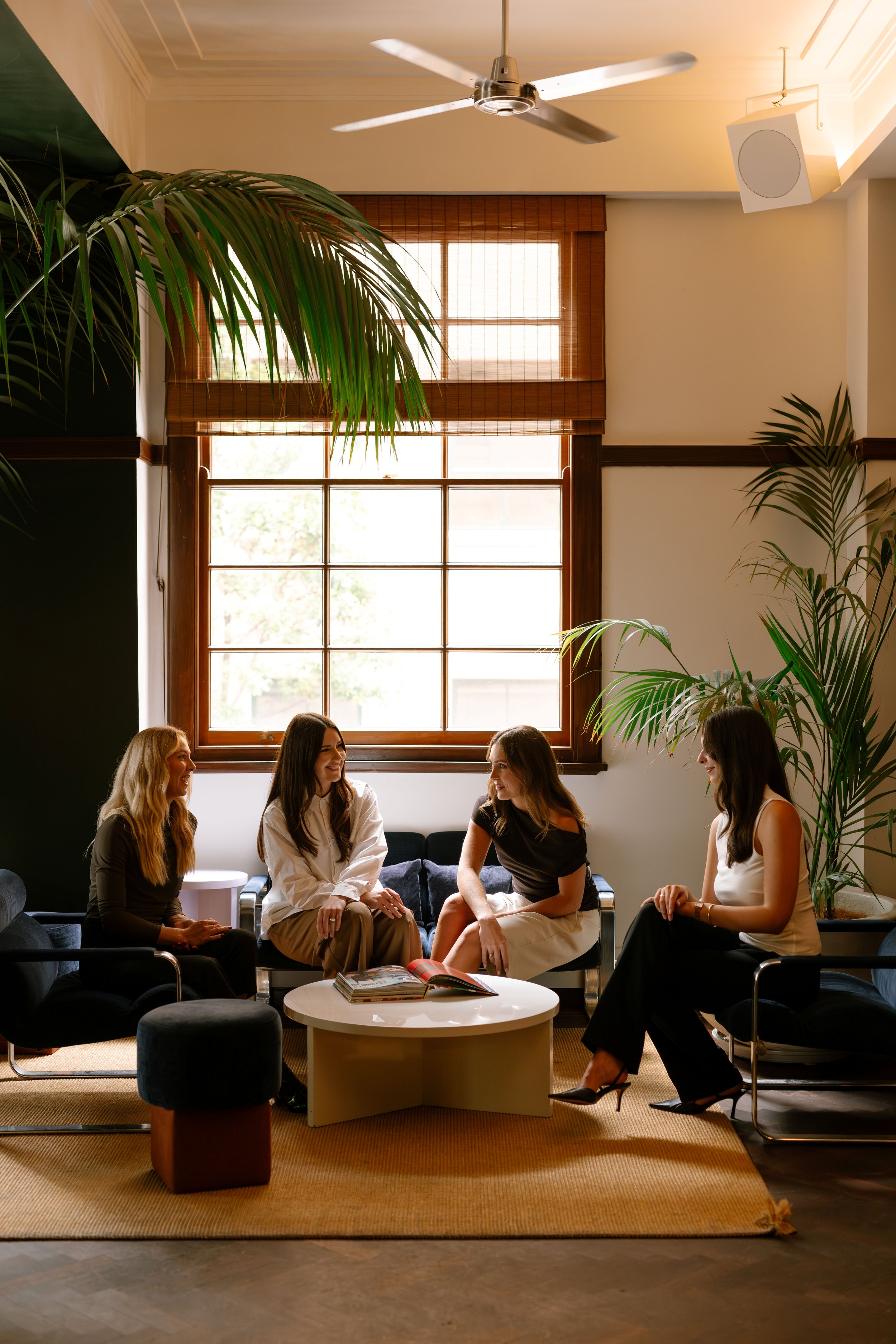 Four women sitting and talking in a modern, well-lit living room with large window, green plants, and a round coffee table with magazines.