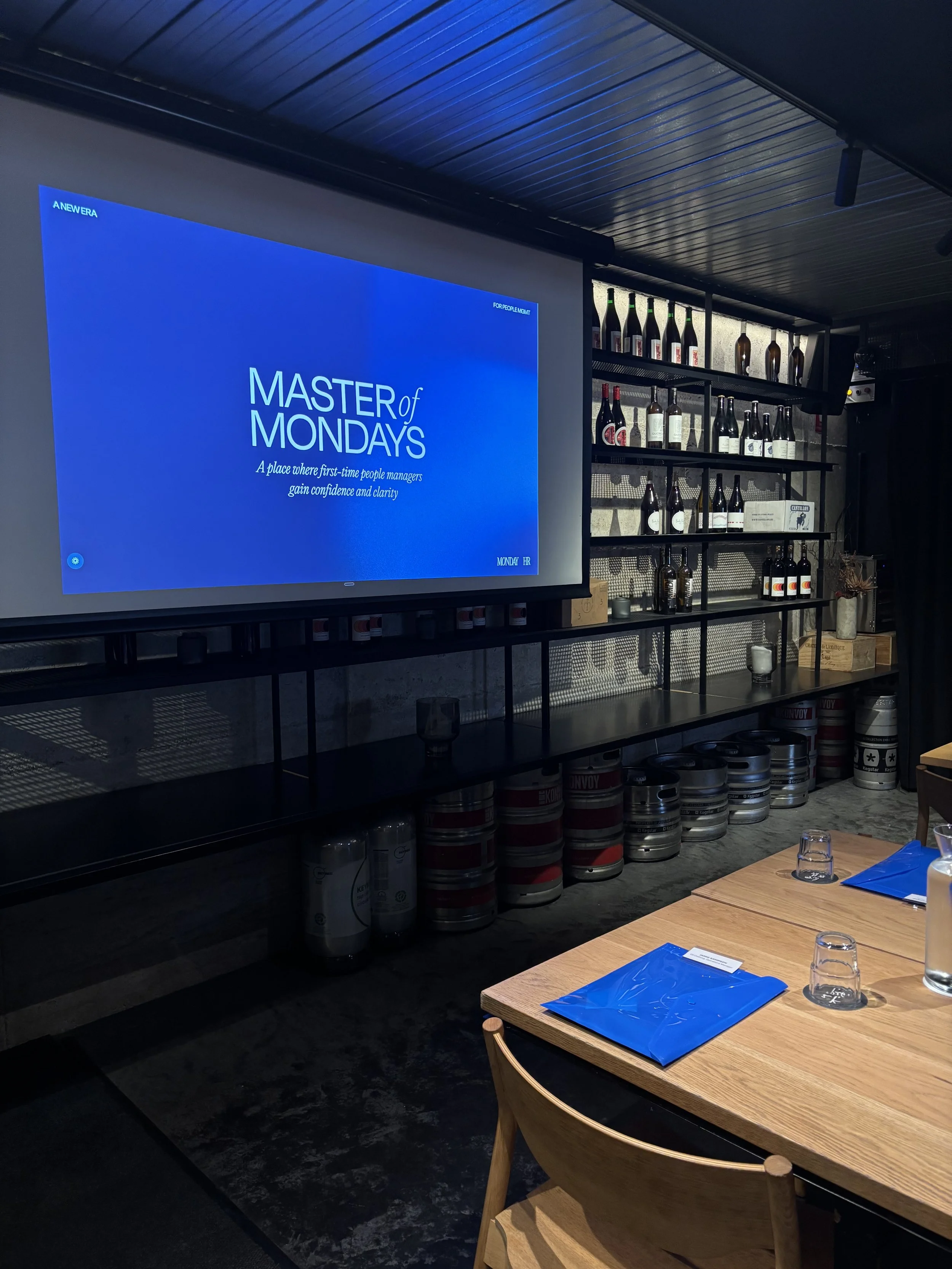 A conference room setup for a training session titled 'Master of Mondays' with a large screen displaying the session title, a table with blue folders, glasses, and water bottles, and a background of shelves with bottles and decorative items.