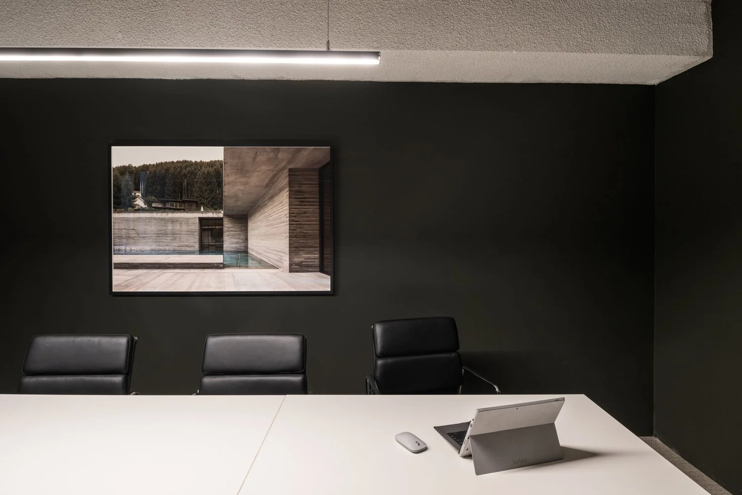 Modern conference room with white table, black chairs, a laptop, mouse, and a large wall art photograph of a contemporary building with wood and concrete design.