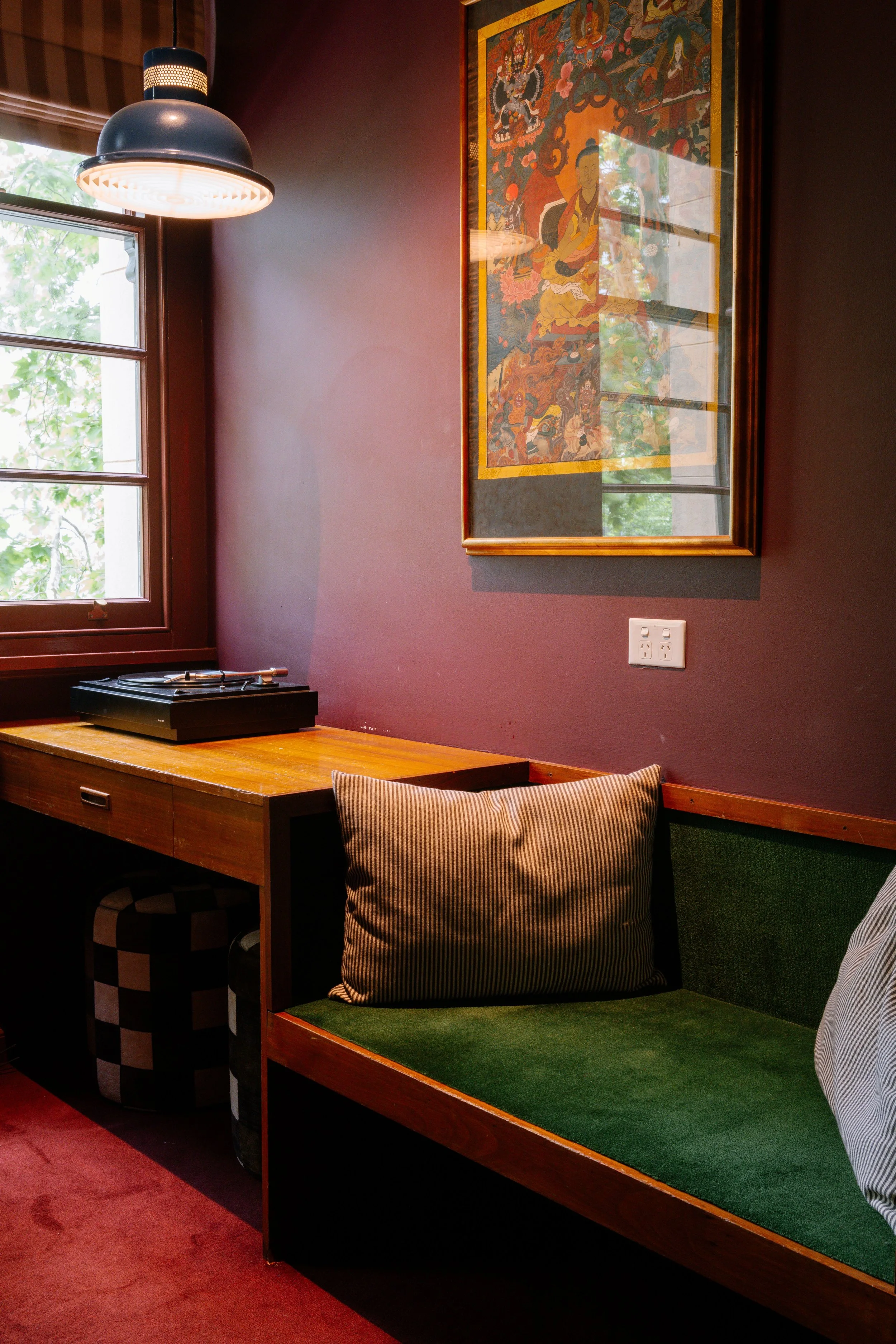 A cozy corner of a room with a wooden desk, a green cushioned bench with striped pillows, a window with blinds, a black pendant light, and colorful artwork hanging on a dark red wall.