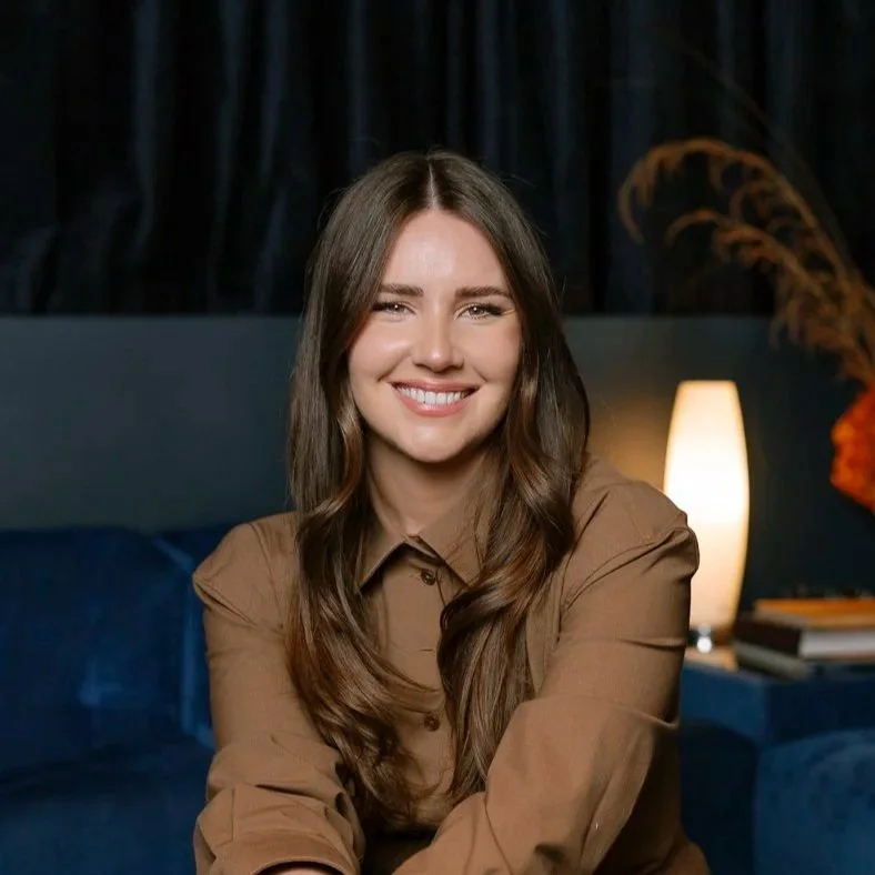Young woman with long brown hair smiling, wearing a brown shirt, sitting on a dark blue sofa in a cozy, dimly lit room with a lamp and decorative plant in the background.