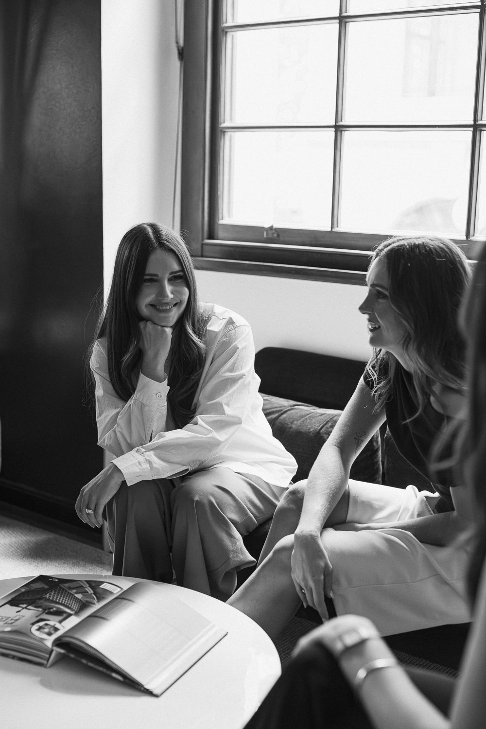 Two young women sitting on a couch, smiling and engaged in conversation, near a window with natural light, with an open magazine on a small table in front of them.