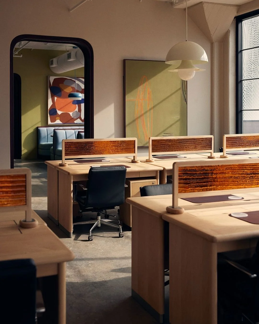 Modern workspace with wooden desks, black office chairs, and natural sunlight coming through large windows. Decor includes abstract art on the walls and a unique white pendant light hanging from the ceiling.