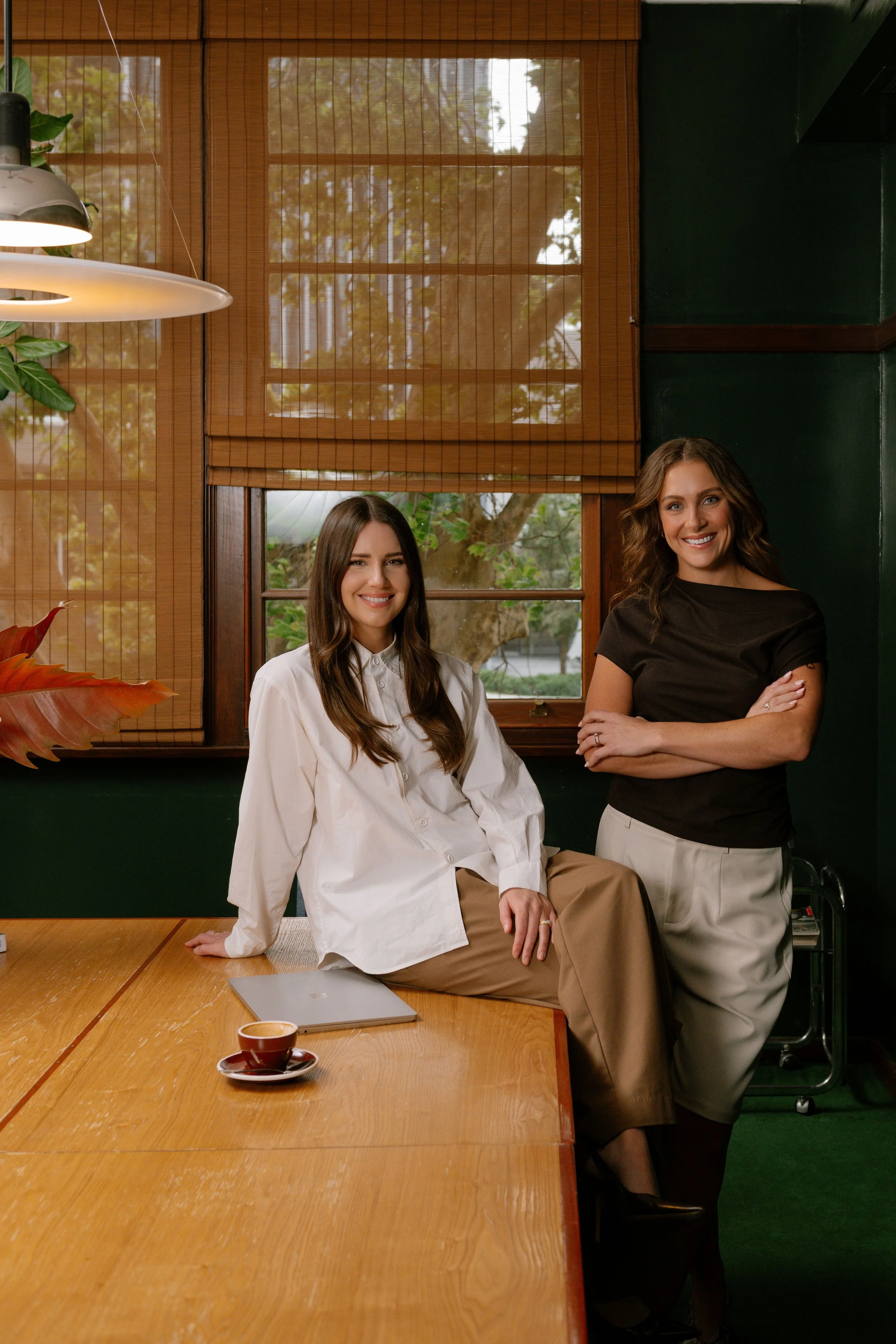 Two women smiling in a cozy room with wooden window blinds, one sitting on a table with a laptop and coffee cup, the other standing with arms crossed.