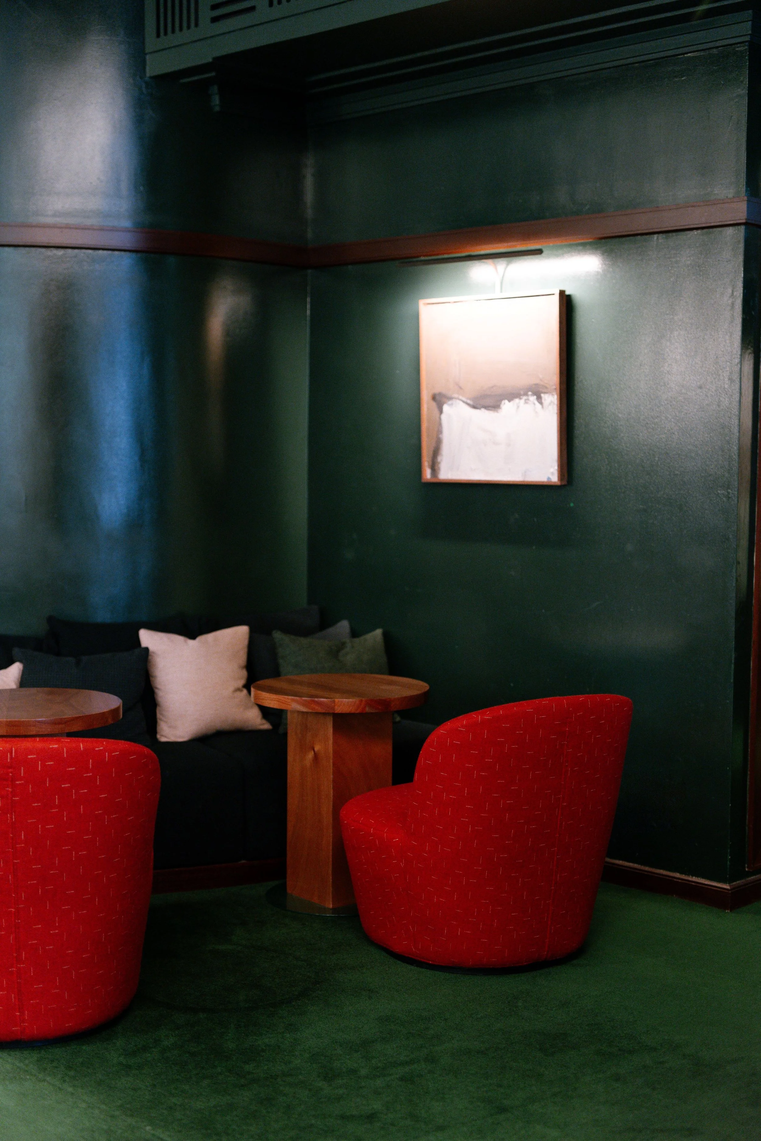 A cozy corner with dark green walls, a black sofa with beige and green cushions, a round wooden table, two red upholstered chairs, a framed abstract painting, and green carpet.