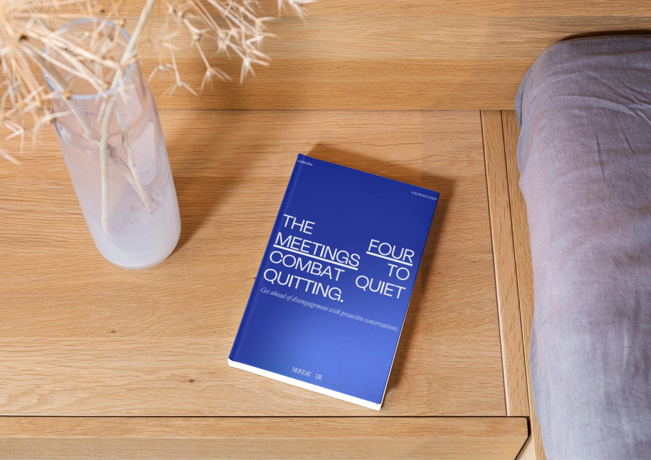 A wooden bedside table with a blue book titled "Four to The Meetings Combat Quiet Quitting" and a clear glass vase with dried branches.