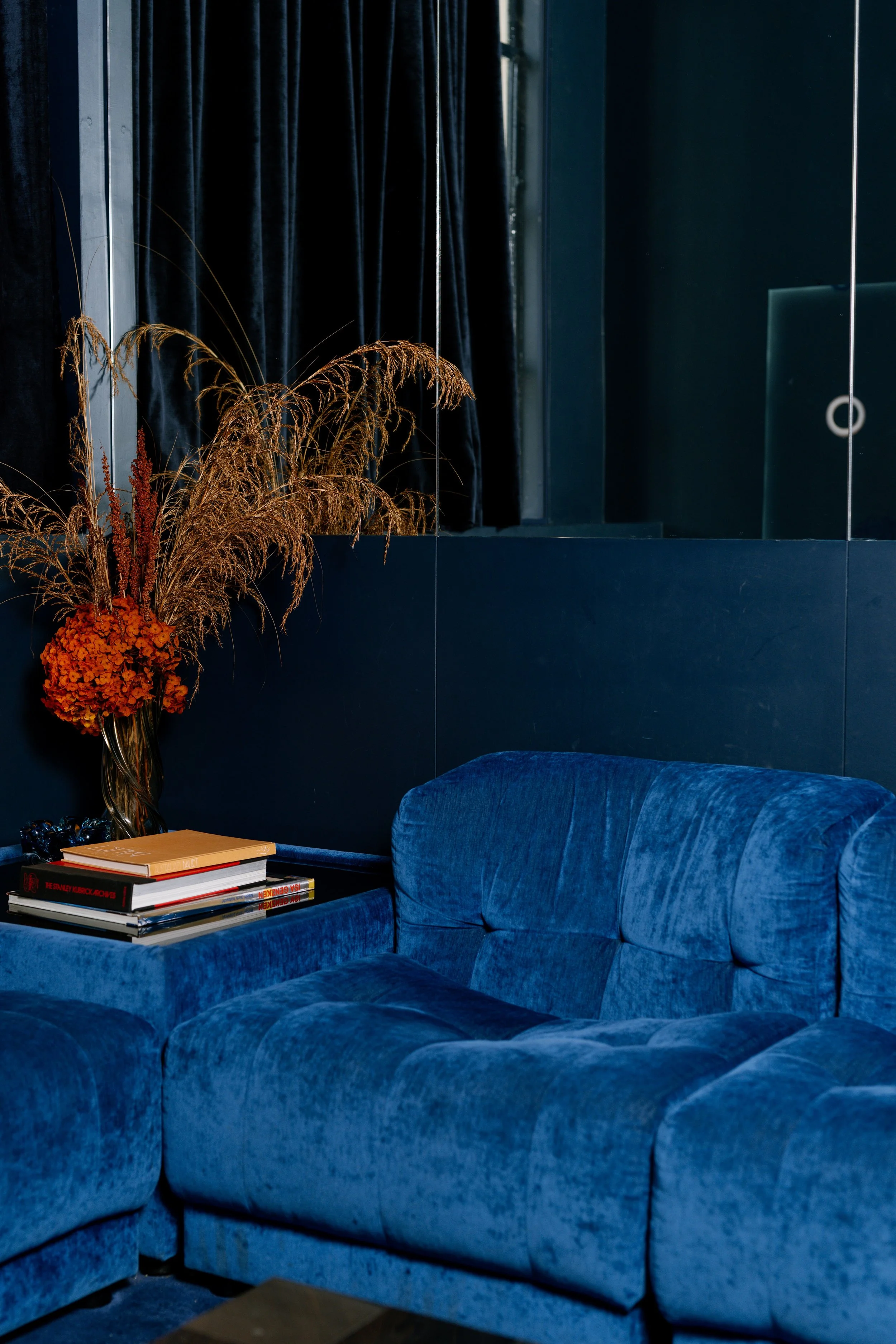 A blue velvet sofa with a side table stacked with books, a vase with dried orange flowers and wheat, dark curtains, and a mirrored wall.