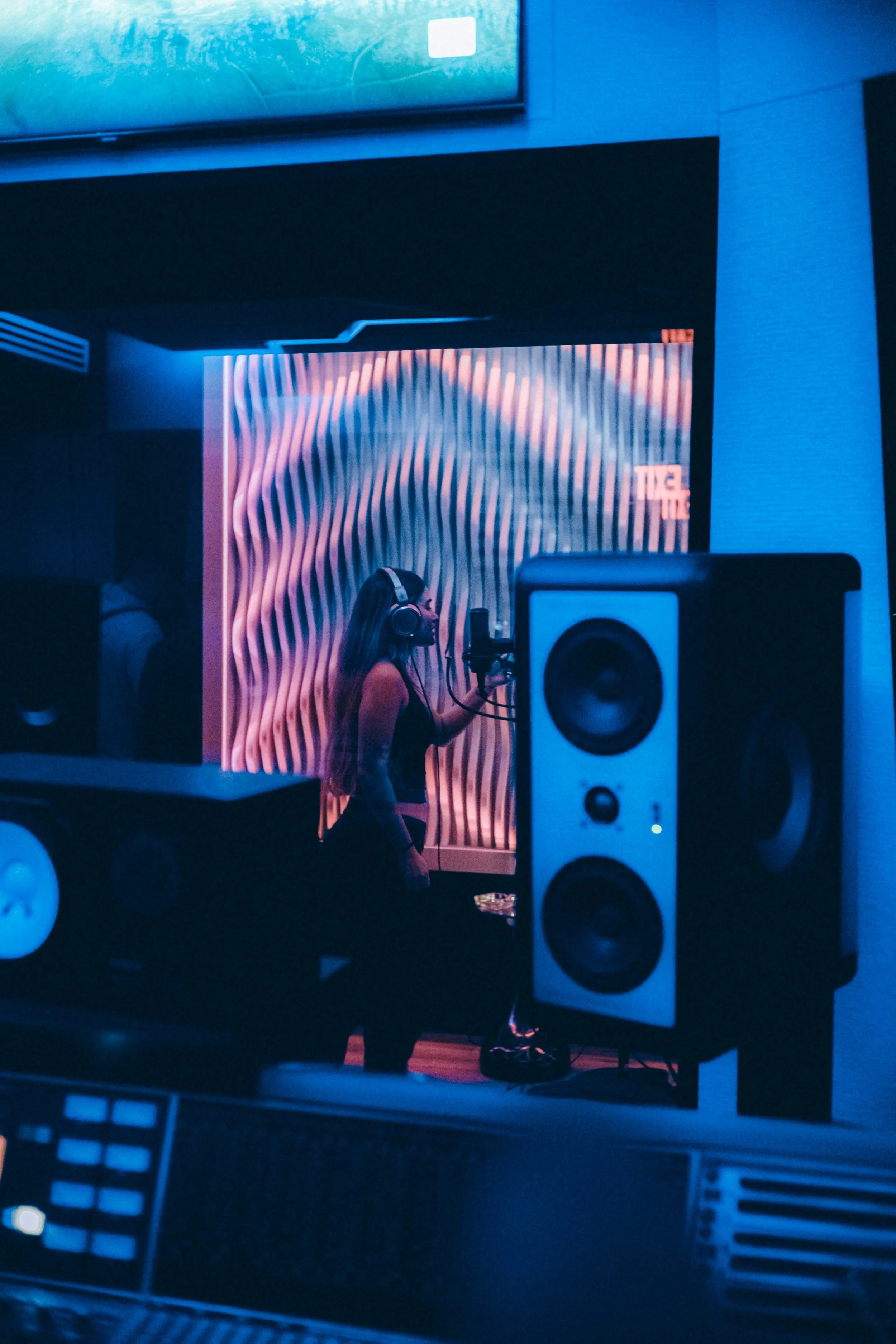 A woman with headphones recording vocals in a music studio with large speakers and colorful, wavy backlit wall.