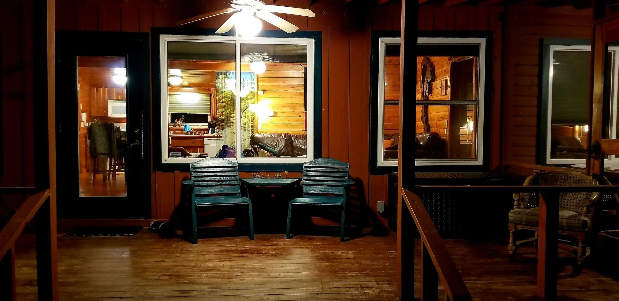 Night view of a wooden porch with two black plastic chairs and a small table, showing the interior of a cozy log cabin with warm lighting, wooden walls, and furniture inside.