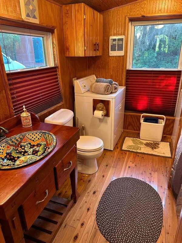 A small bathroom with wooden walls and flooring, featuring a wooden vanity with a colorful decorative sink, a toilet, a washing machine with towels on top, two windows with wooden blinds, a trash bin, a patterned rug, and a black and white oval rug.