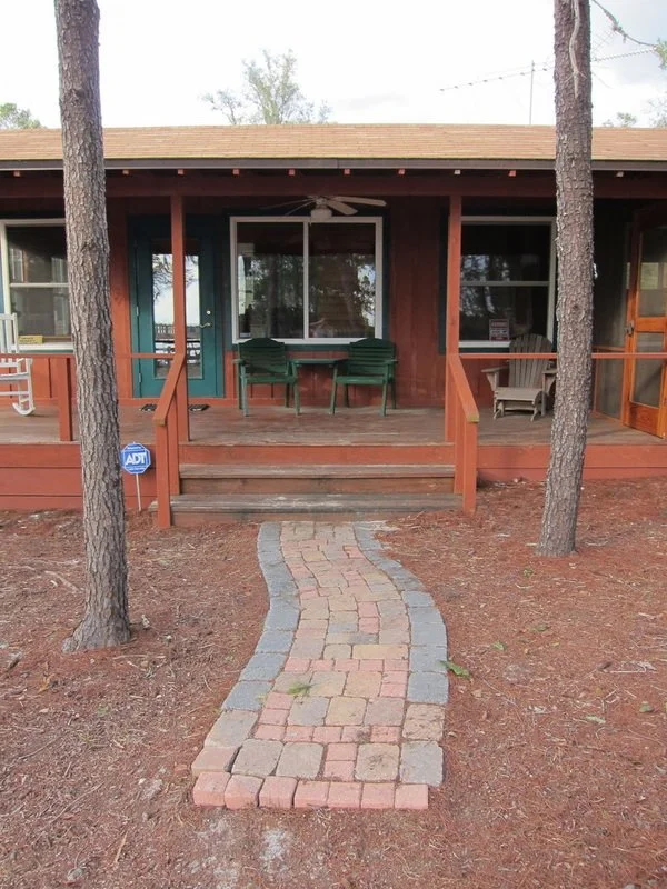 A house with a front porch, a small brick pathway leading to it, outdoor chairs, and trees in the yard.