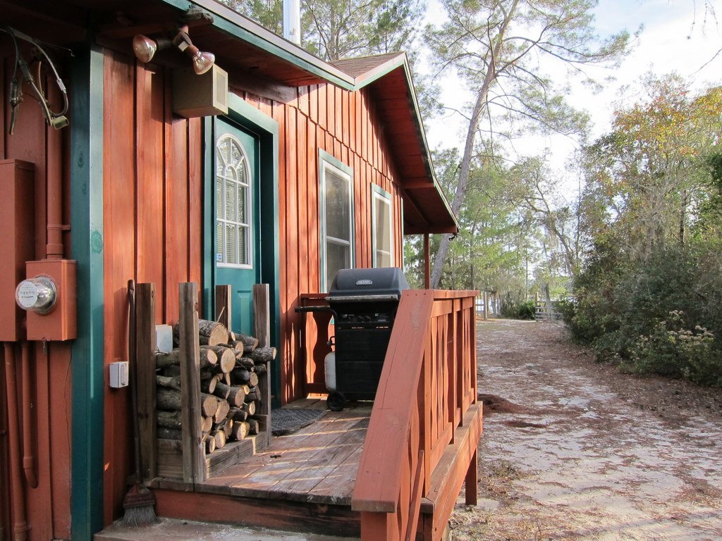 Red wooden house with a small porch, firewood stacked against the wall, a grill on the porch, and trees in the background.