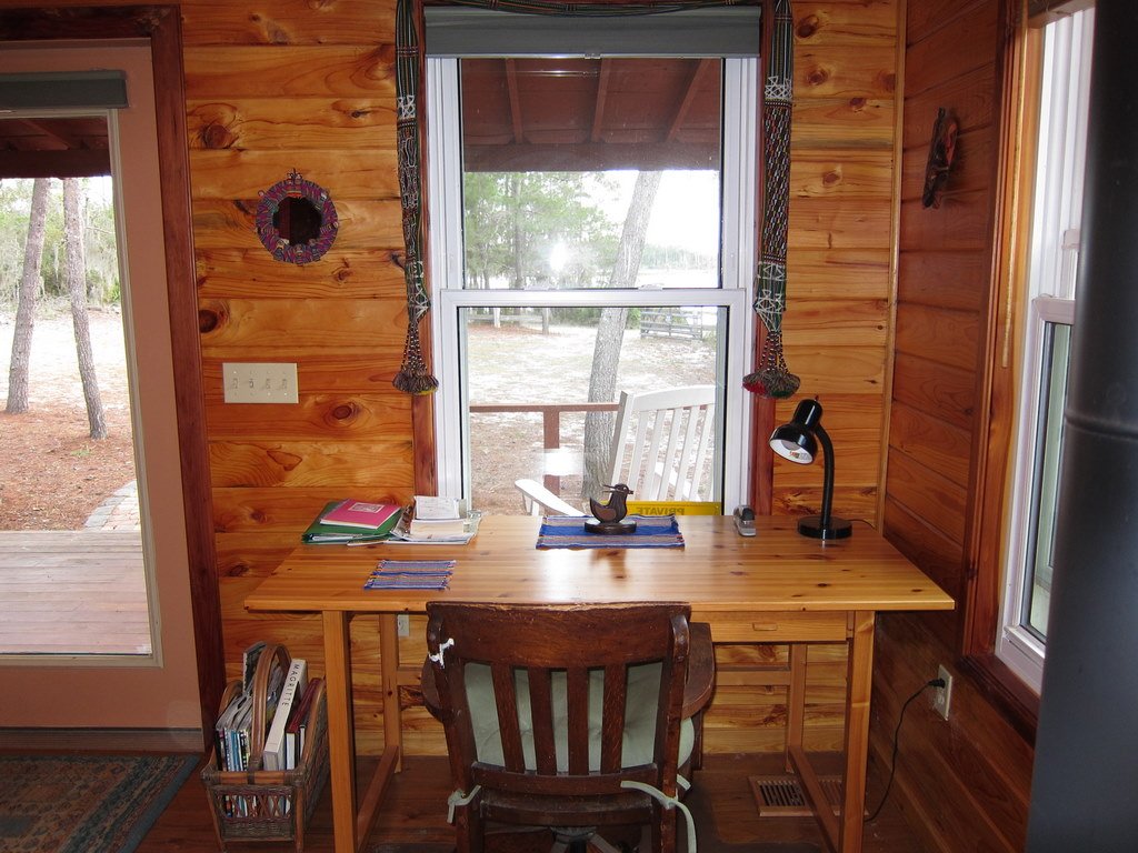 Small wooden desk with a lamp, papers, and a teapot, placed by a window inside a wooden cabin, overlooking trees outside.