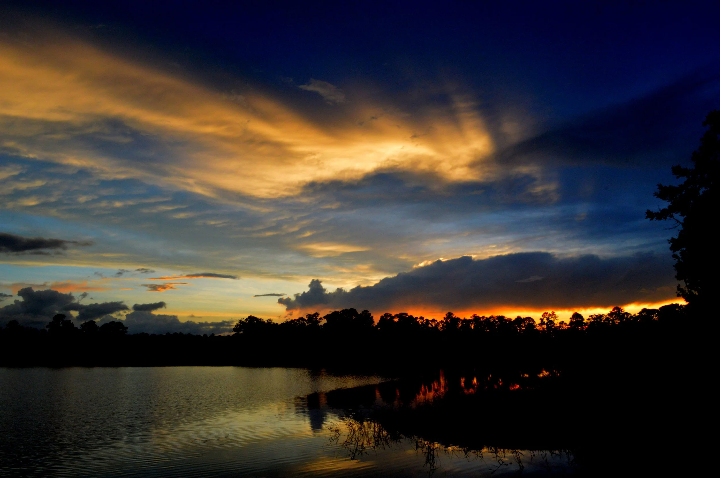 Sunset over a lake with dark silhouettes of trees along the shoreline, colorful clouds in the sky, and reflected light on the water.