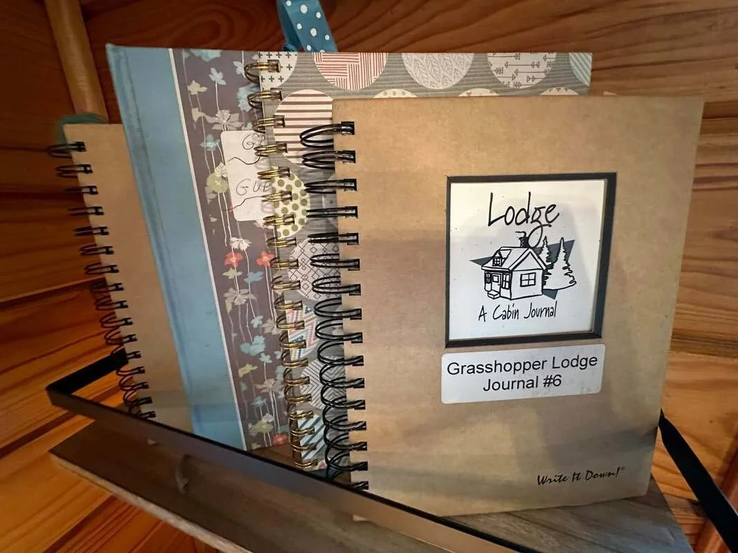 Open cabin journal titled 'Lodge: A Cabin Journal, Grasshopper Lodge Journal #6' with a drawing of a cabin and trees on the cover, inside a wooden cabin.