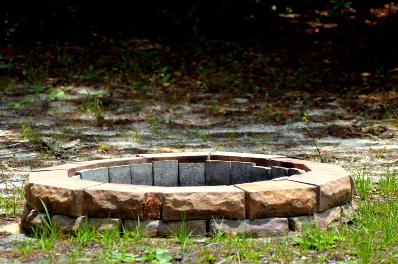 A circular fire pit made of bricks and stones on grass and dirt ground in an outdoor setting.