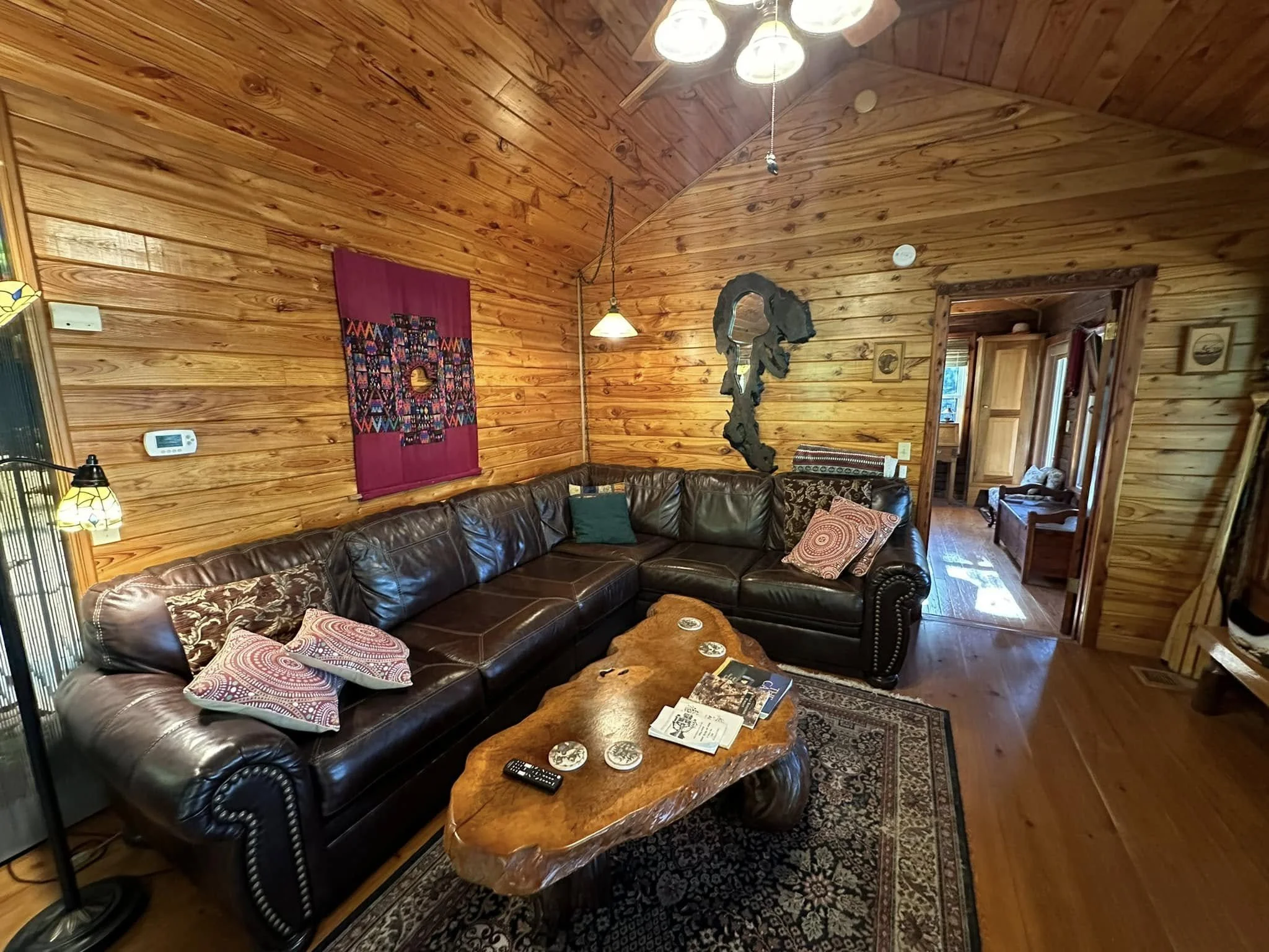 Wood-paneled living room with a large dark leather sectional sofa, a rustic wooden coffee table, wall art, hanging lamps, and an entrance to another room with wooden furniture.