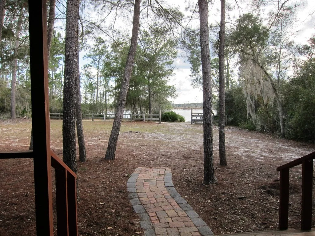 View from a porch looking out over a wooded area with a river beyond, a brick pathway, and a wooden fence near the water.