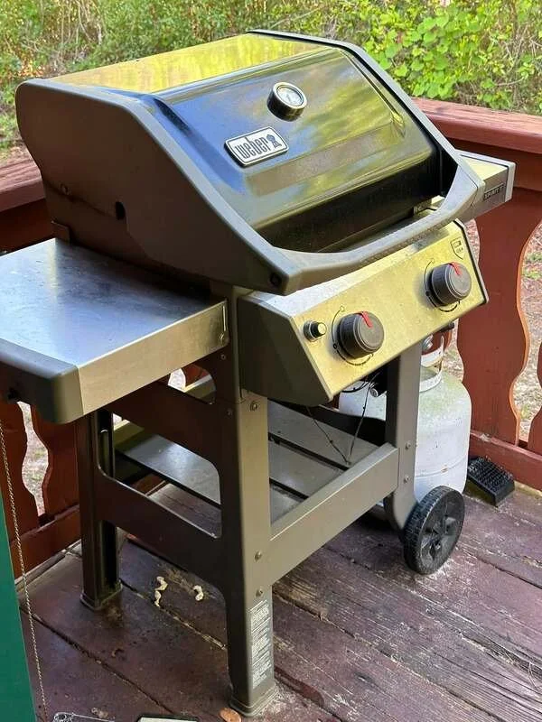 Black and gray Weber gas barbecue grill on a wooden deck with a red railing and green foliage in the background.