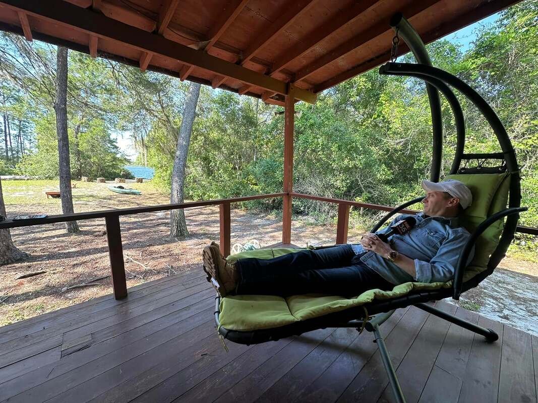 A man relaxing on a green cushioned outdoor lounge chair on a wooden porch, surrounded by trees and greenery.
