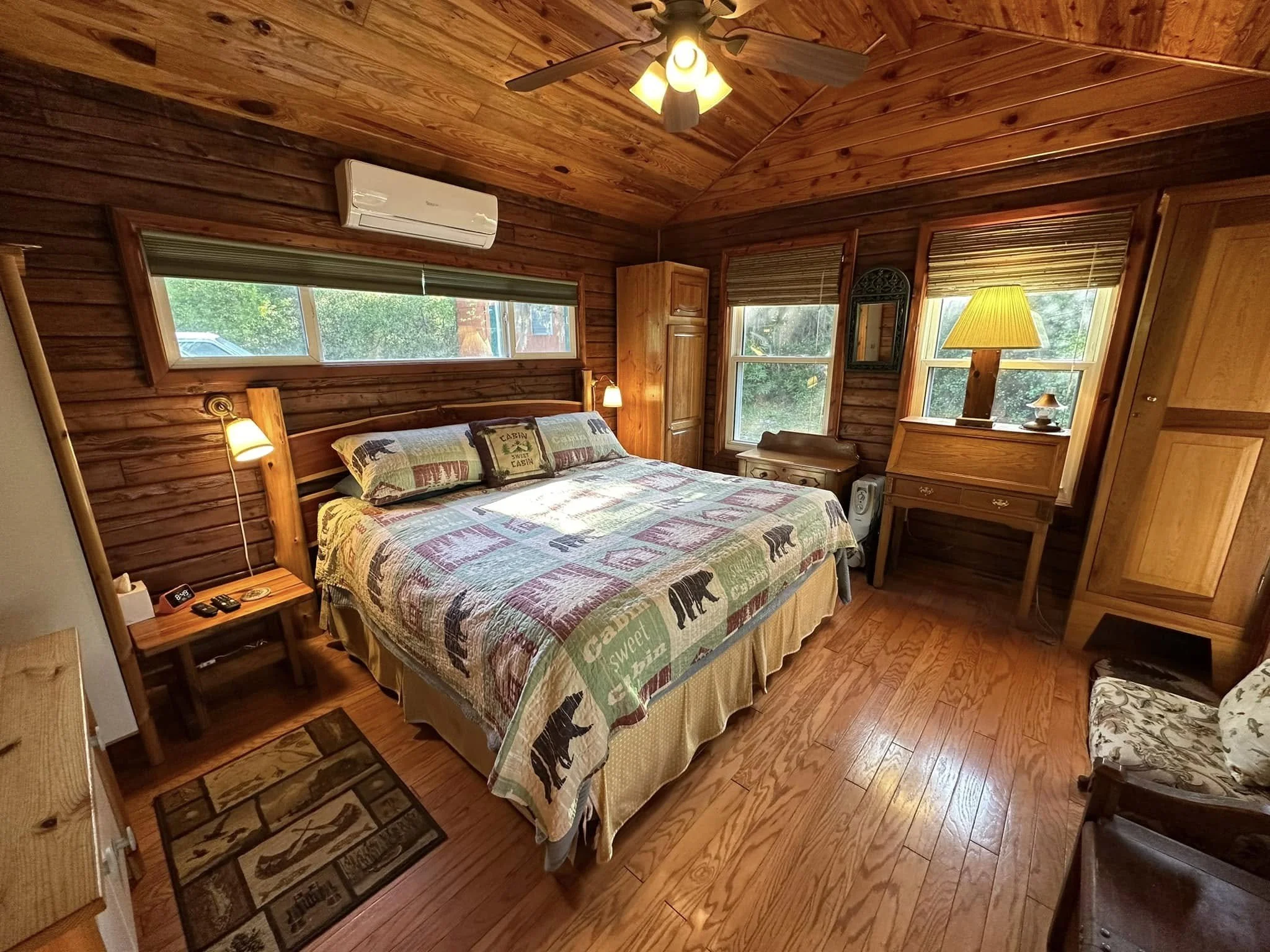 A cozy bedroom with wooden walls, ceiling, and flooring. Features a large bed with a bear-themed quilt, bedside lamps, a window air conditioning unit, and multiple windows with blinds letting in natural light. There is a small wooden dresser, an armc