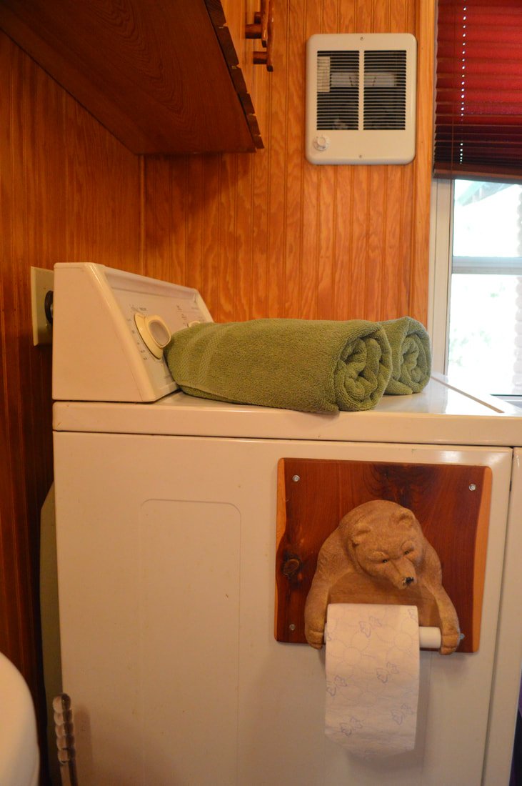 A top-loading washing machine with two rolled green towels on top, mounted on a wooden wall with a vent and window in the background.