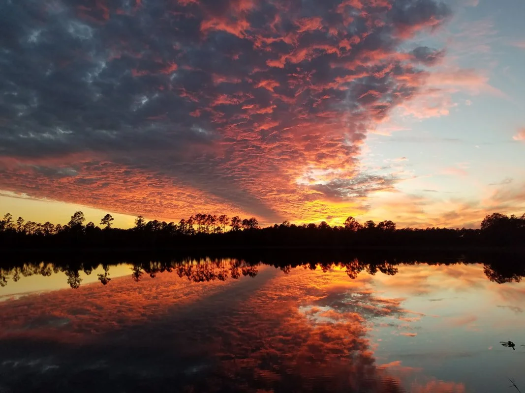 A vibrant sunset over a lake with colorful clouds reflected on the water, and silhouettes of trees along the horizon.