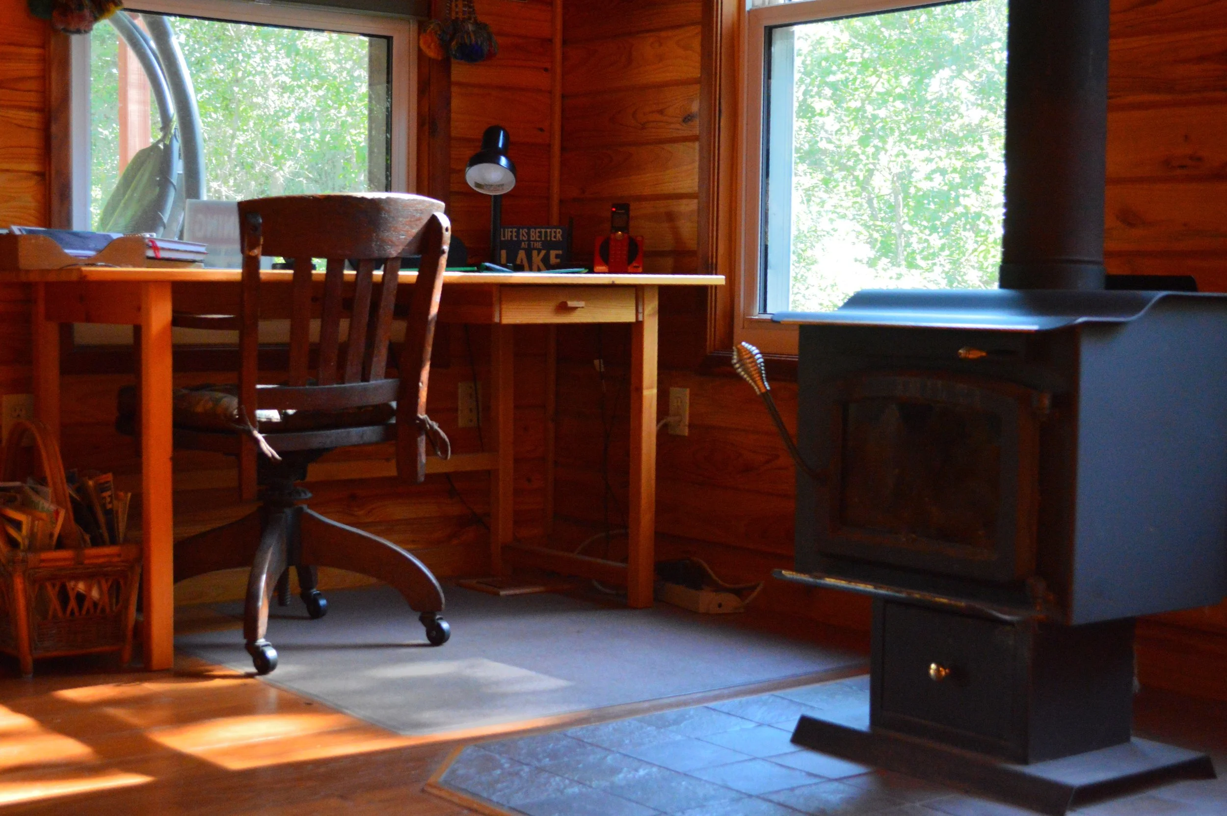 Interior of a cozy wood-paneled room with a wooden desk and chair, a small black stove, and two large windows showing green trees outside.