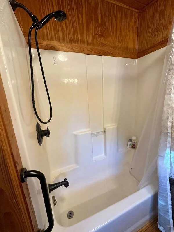 Empty shower with a wooden panel ceiling, white walls, a built-in seat, a handheld showerhead, and bottles of shampoo or soap on the corner shelf.
