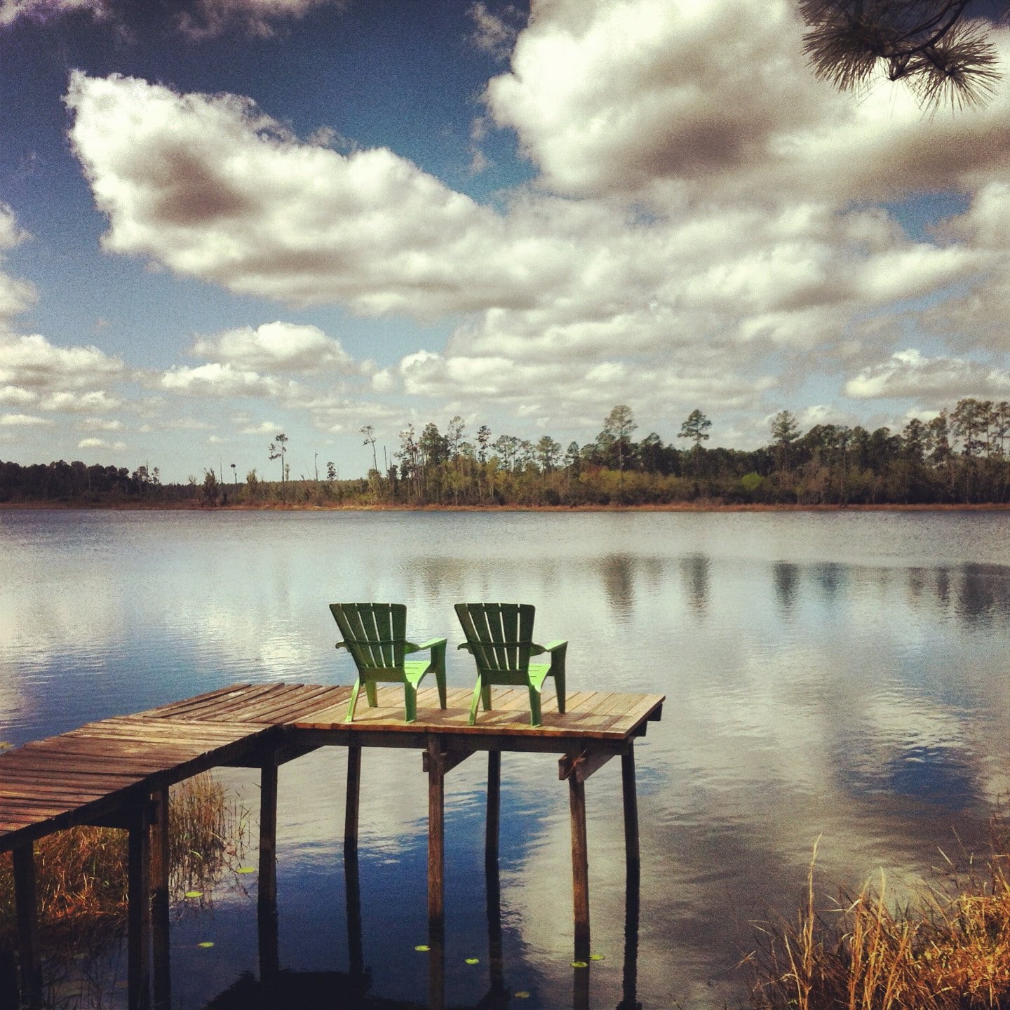 A lakeside scene with a wooden dock extending into the water, two green chairs on the dock, and a cloudy sky reflected on the calm lake surface, with trees in the distance.
