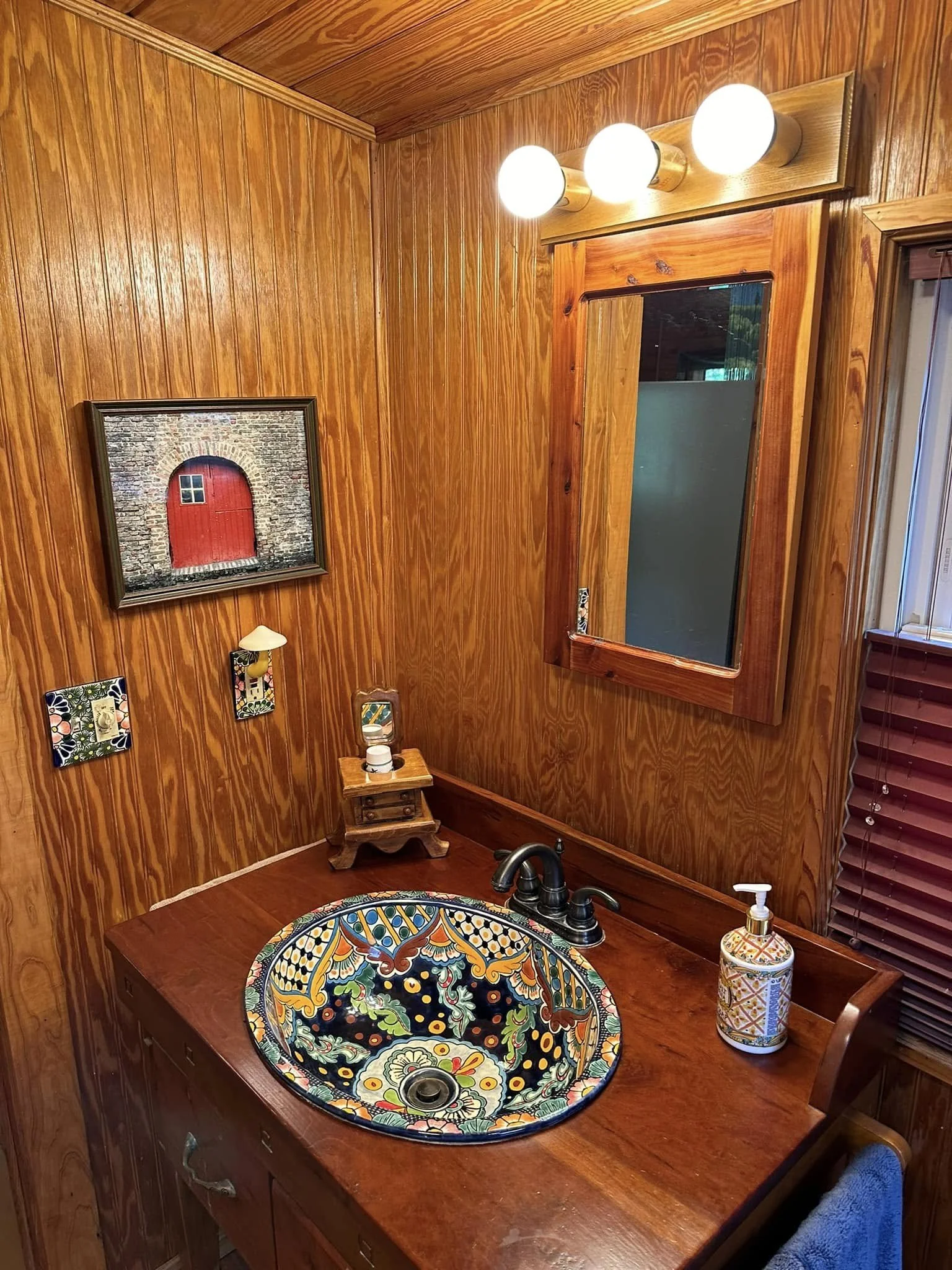 Wood-paneled bathroom with a colorful decorative sink, a wooden framed mirror, a three-bulb light fixture, a framed picture of a red door, a soap dispenser, and a towel hanging on a hook.