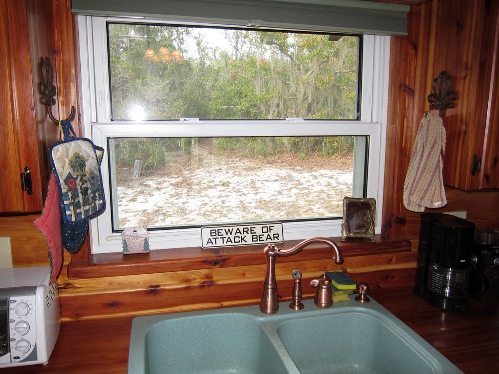 Kitchen window with a wooden frame, showing a wooded outdoor area. Inside, on the window sill are a sign that reads "Beware of Attack Bear," a small house-shaped ornament, and a silver-colored tray. The kitchen sink with a bronze faucet is below the 