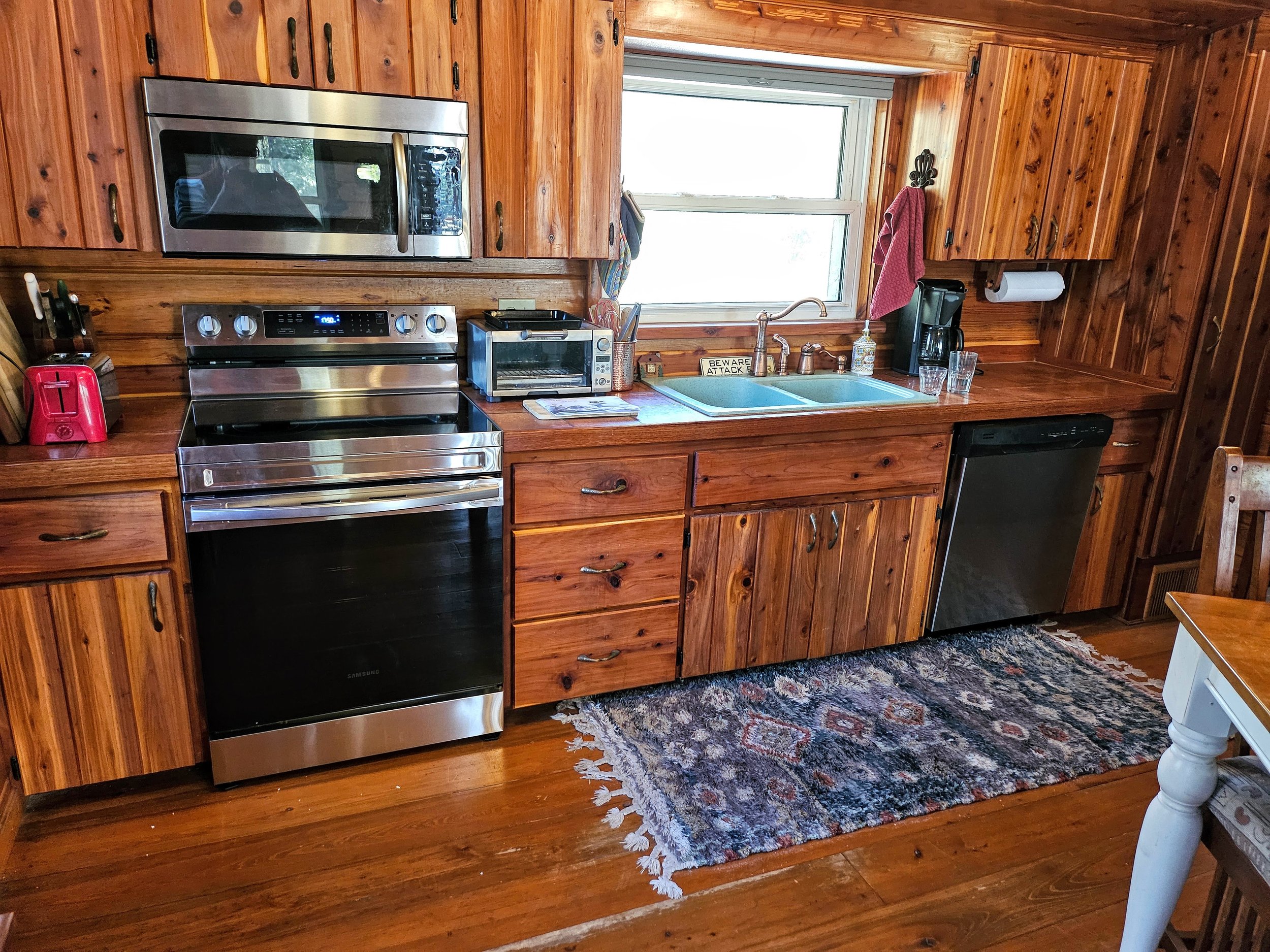 A rustic kitchen with wood-paneled walls and cabinetry, stainless steel appliances including a microwave, stove, and dishwasher, a window above the sink, and various small appliances on the countertop, with a patterned rug on the wooden floor.