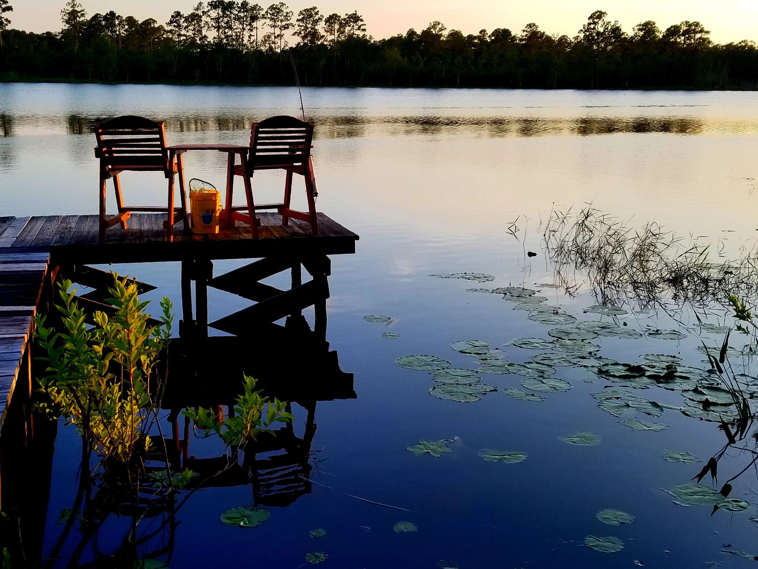 A lakeside scene at sunset with two wooden chairs and a small table on a dock, a bucket, and aquatic plants on the water.