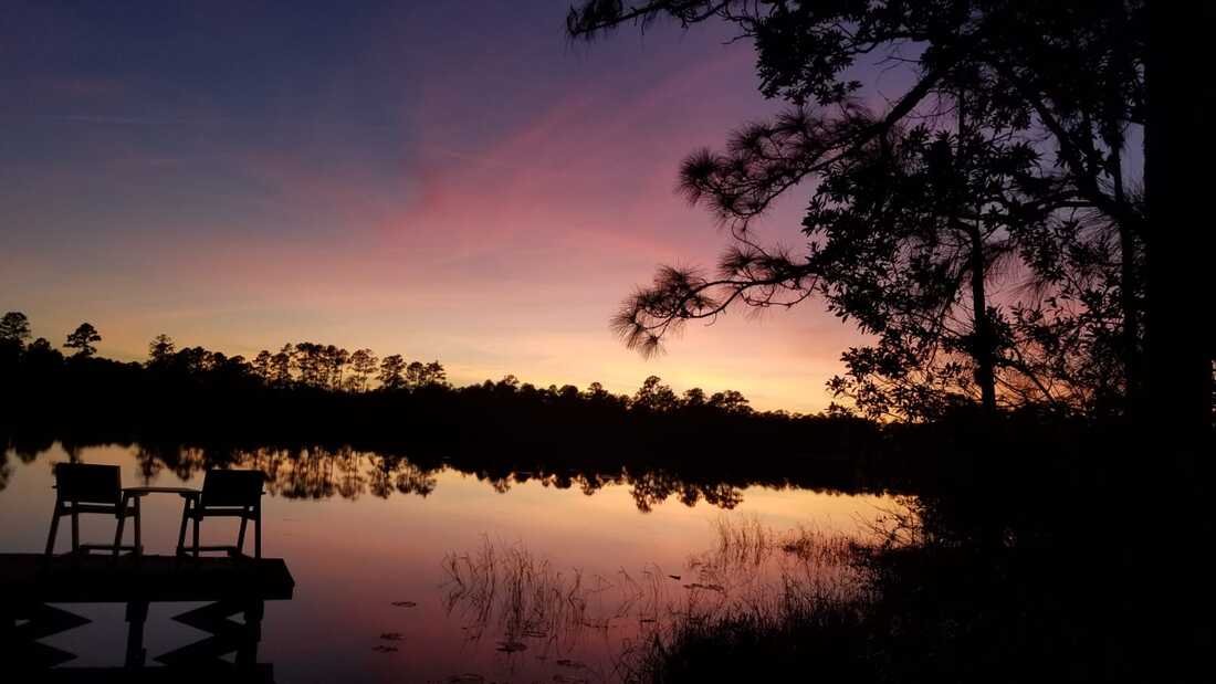 Silhouette of two chairs on a dock beside a lake during sunset, with colorful sky and trees reflected in the water.
