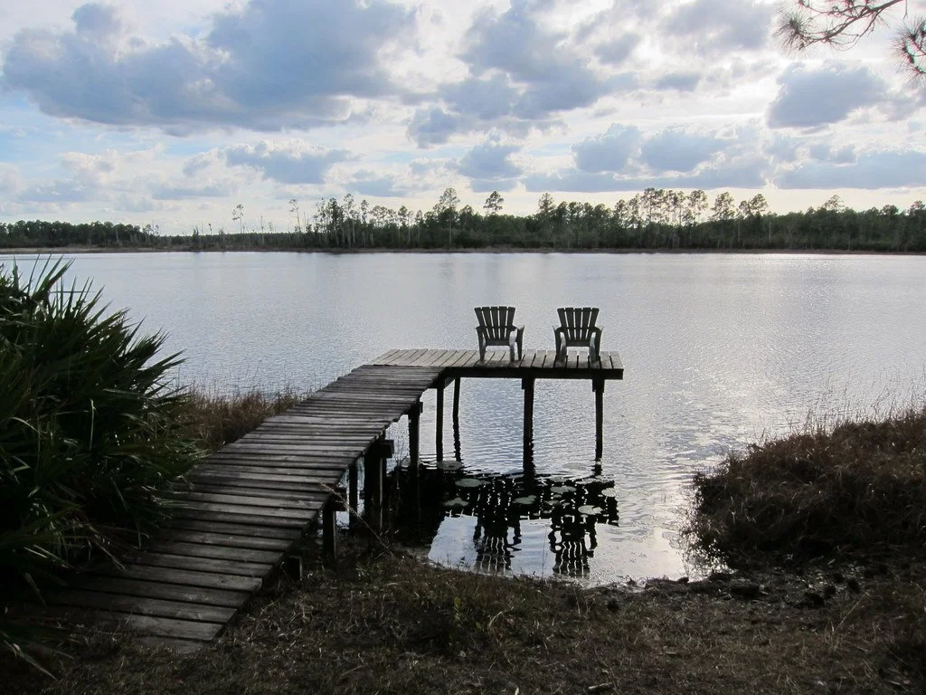 A wooden dock with two chairs extends over a calm lake under a partly cloudy sky.