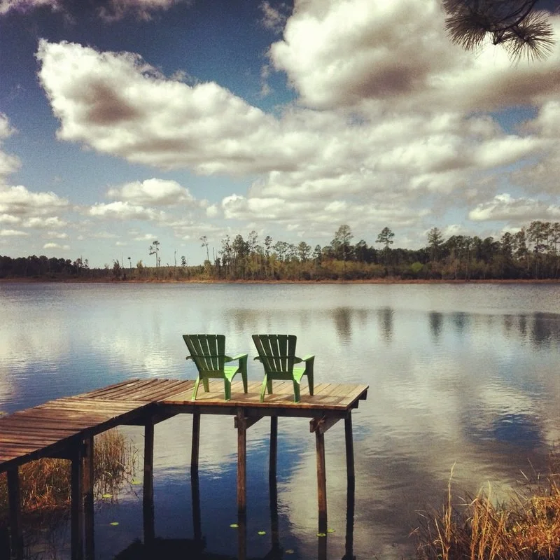 Two green Adirondack chairs on a wooden dock overlooking a calm lake with a treeline in the distance and a partly cloudy sky.