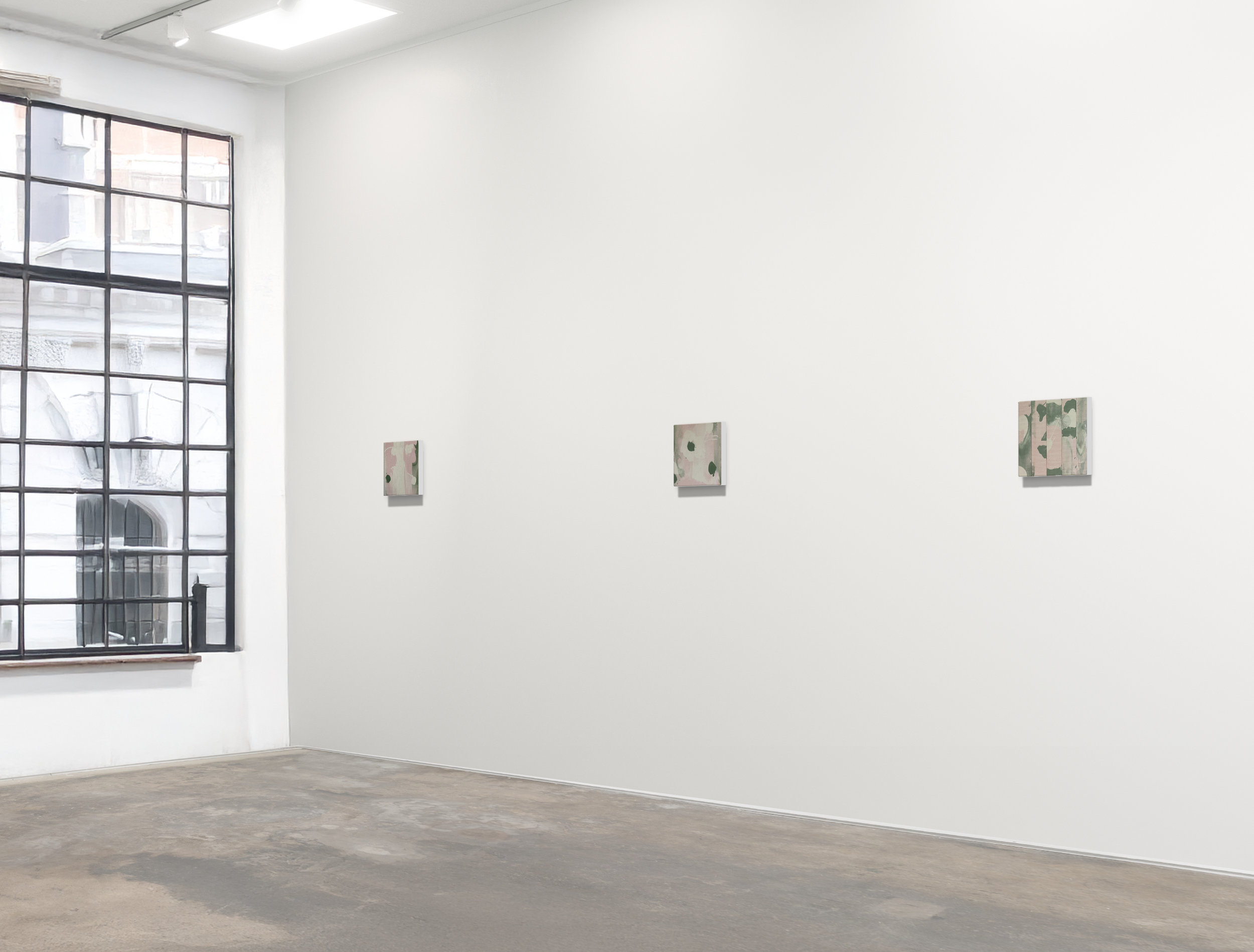 Art gallery with three small abstract paintings on a white wall, large window with metal grid on the left, concrete floor.
