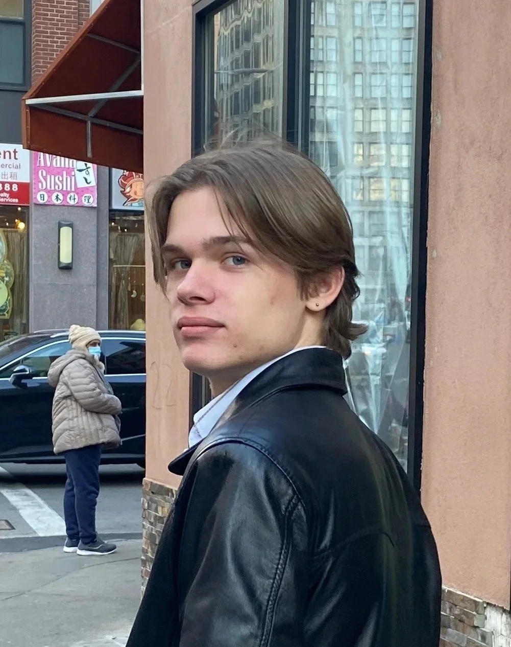 A young man with brown hair and blue eyes looking back over his shoulder on a city street, wearing a black leather jacket and a white shirt. In the background, a woman in a beige jacket and face mask stands near a parked car, with store signs and tall buildings reflecting in the glass window behind him.