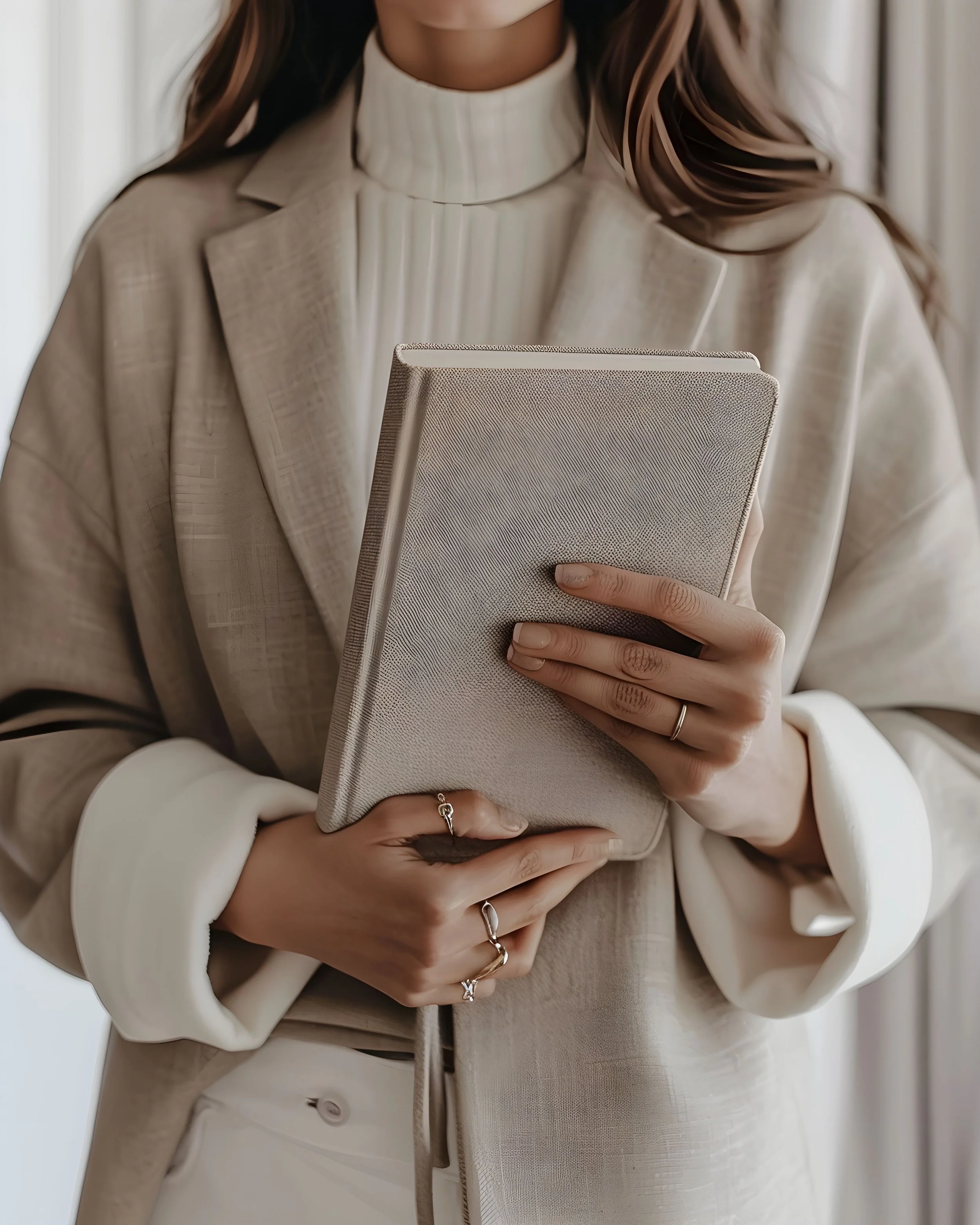 A woman wearing a beige blazer and a white turtleneck, holding a light-colored textured notebook, with her hands adorned with rings.