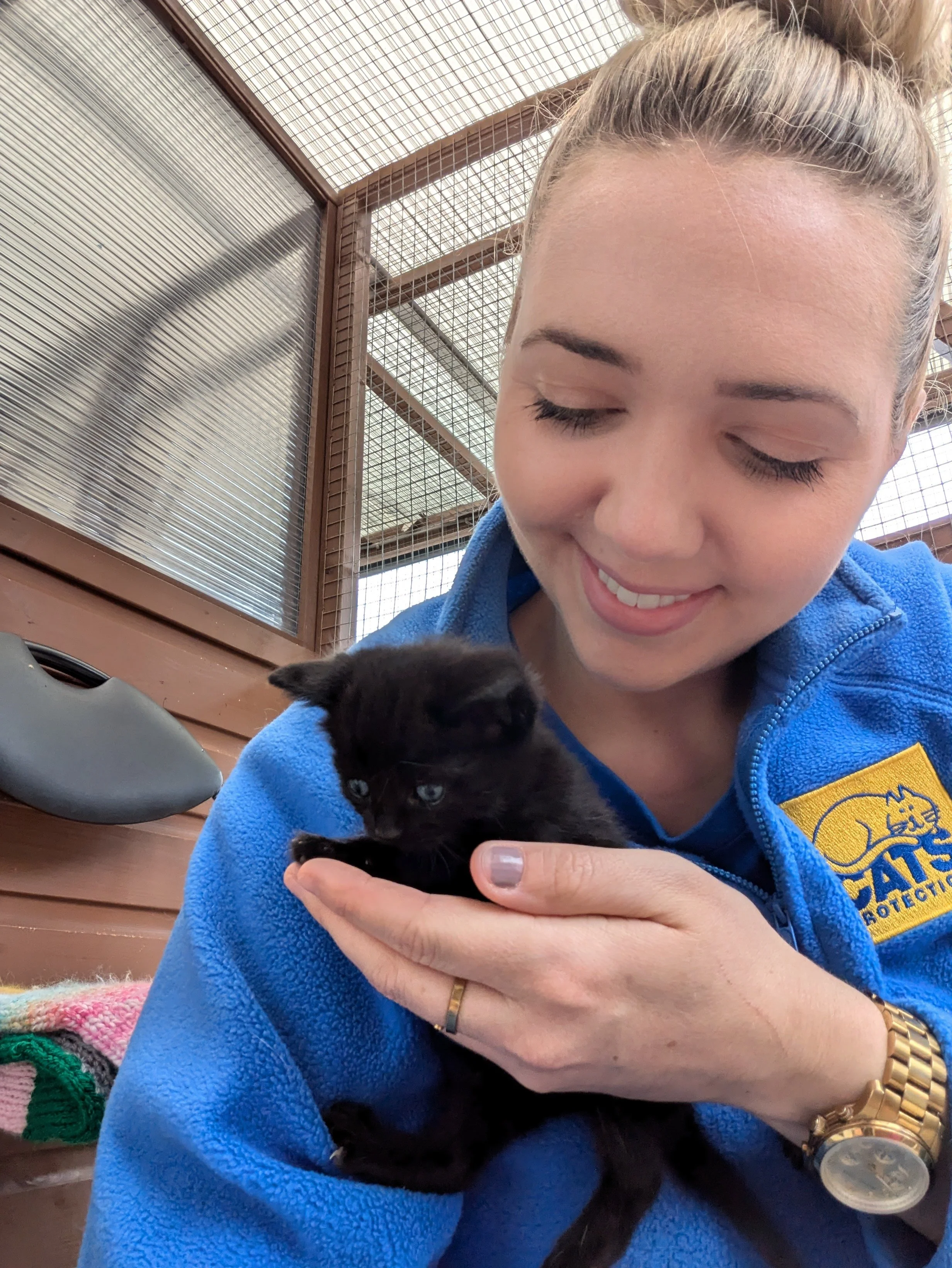 A smiling woman in a blue fleece Cats Protection jacket holding a small black kitten in her hand, inside an outdoor cage.