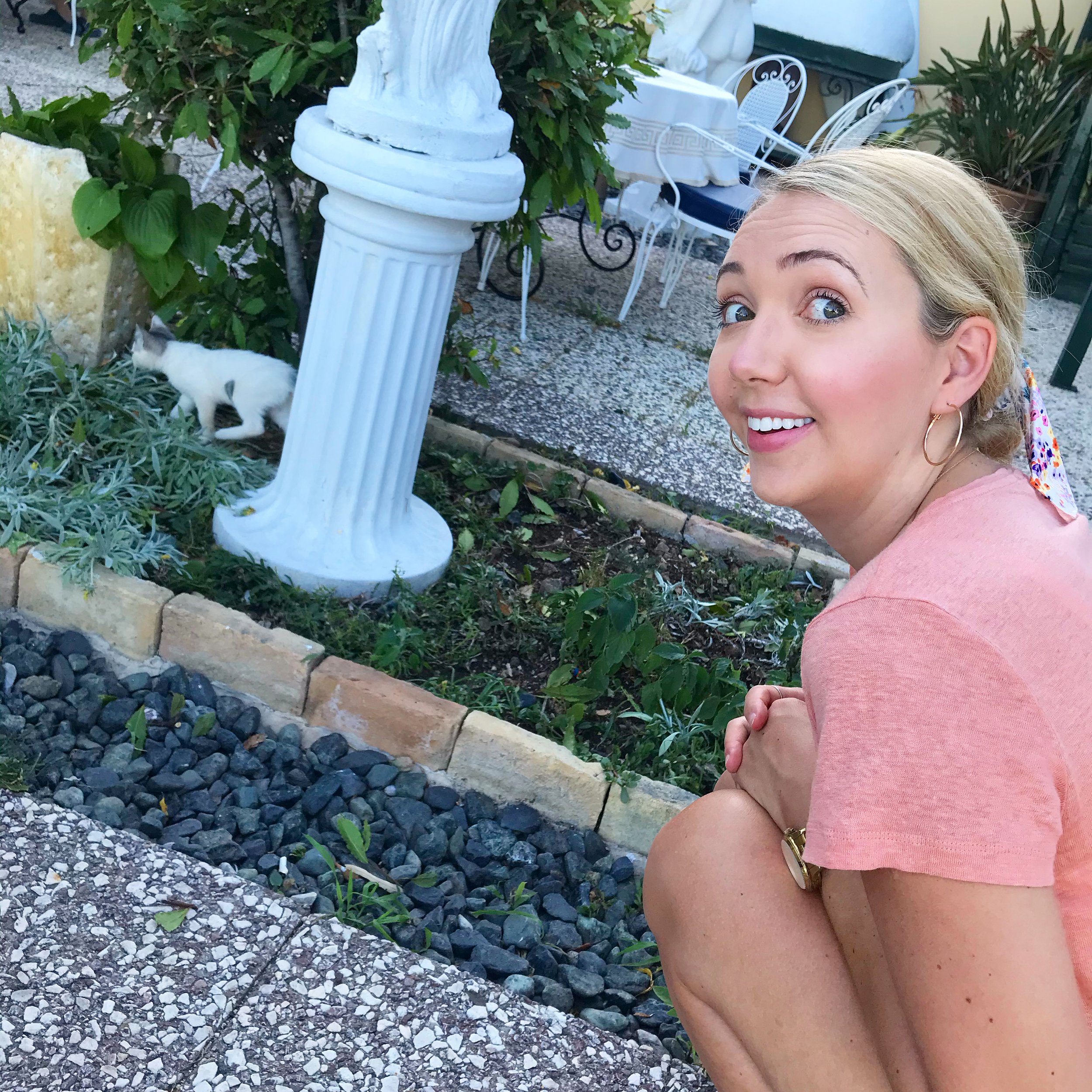 Christie crouching outdoors and smiling at a tiny kitten near garden plants