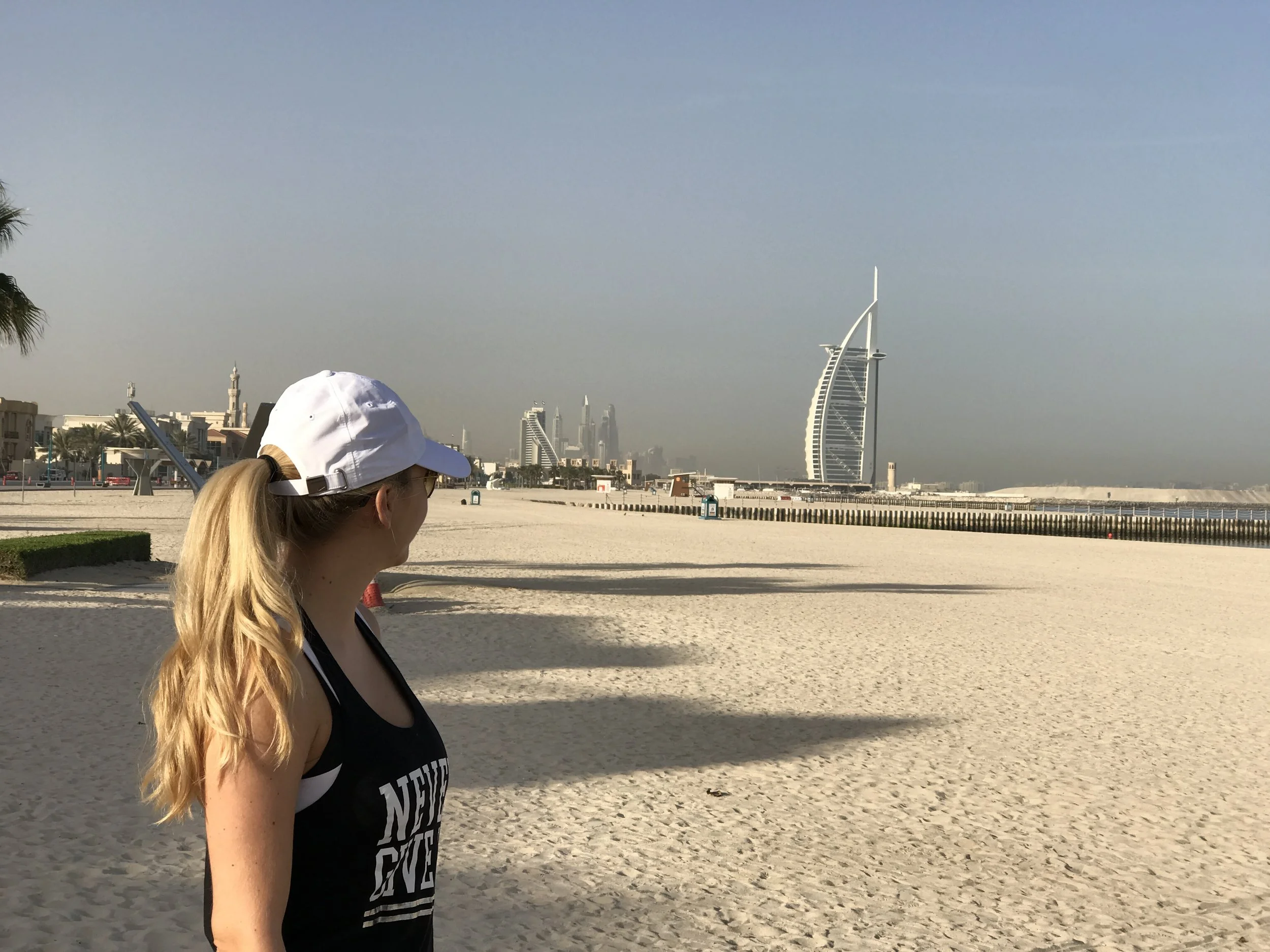 A woman with blonde hair in a ponytail, wearing a white cap and a black tank top with white text, stands on a sandy beach looking towards the city skyline in Dubai, with the Burj Al Arab hotel visible in the distance.