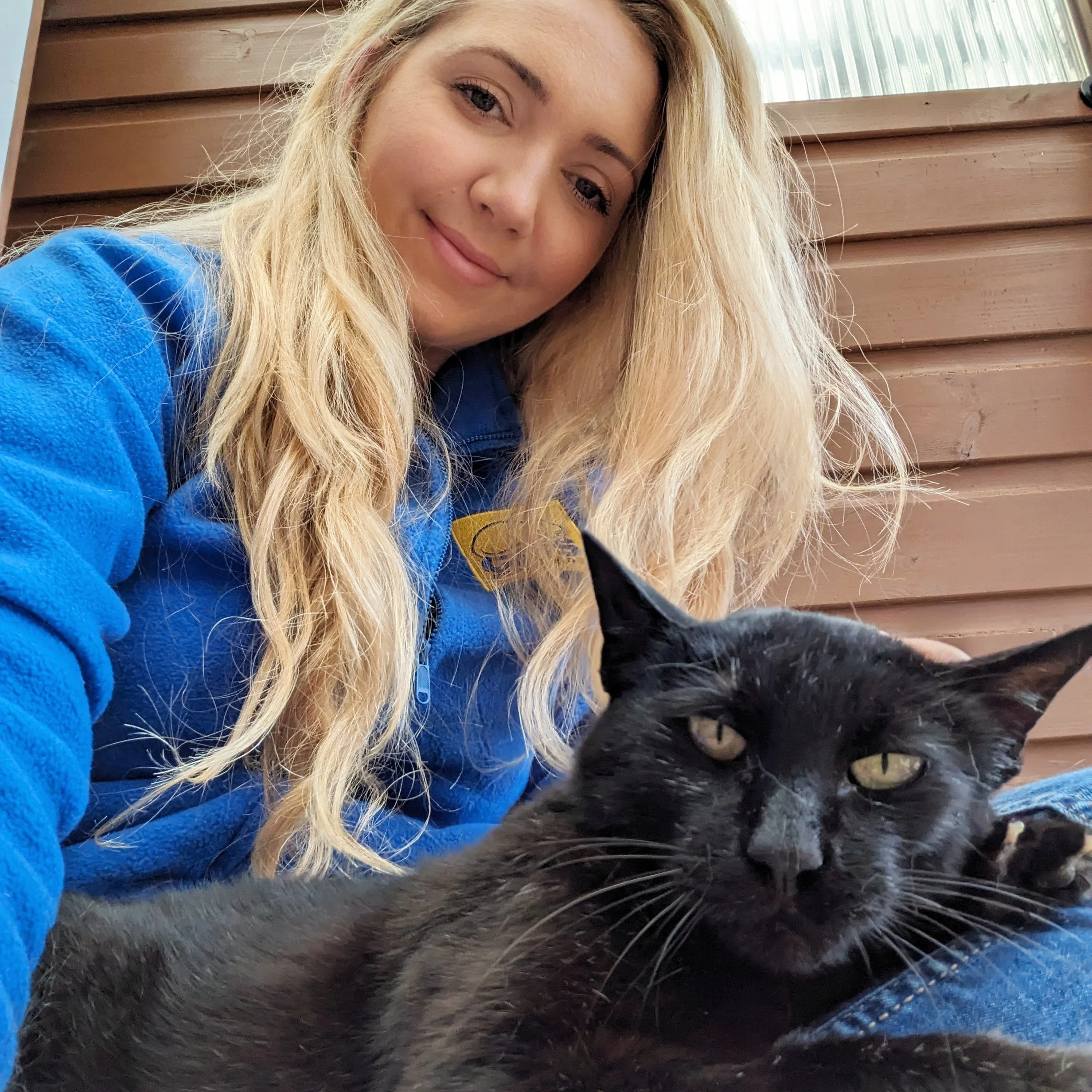 A young woman with long blonde hair and a blue Cats protection jacket, smiling while lying next to a black cat with yellow-green eyes, against a wooden wall background.